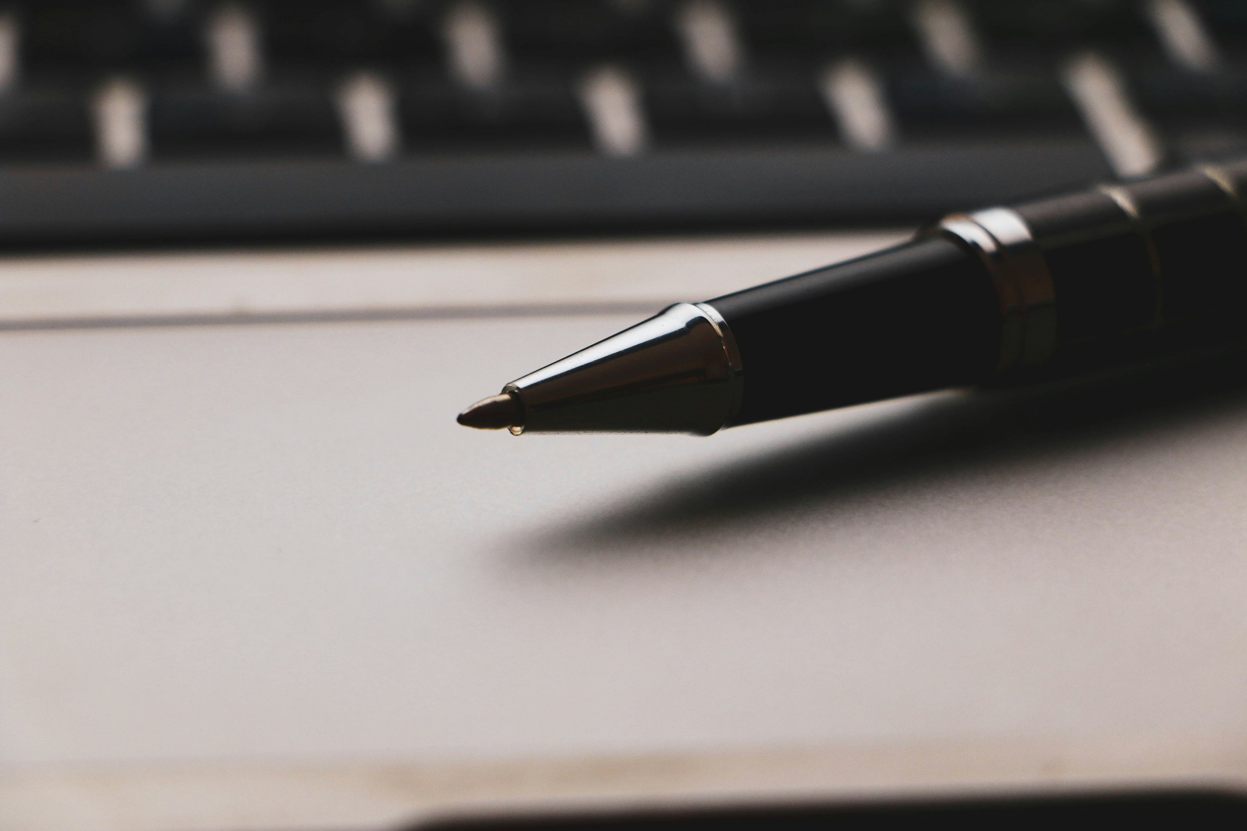 Close-up of a black pen with a silver clip resting on a white surface, with a blurred black keyboard in the background.