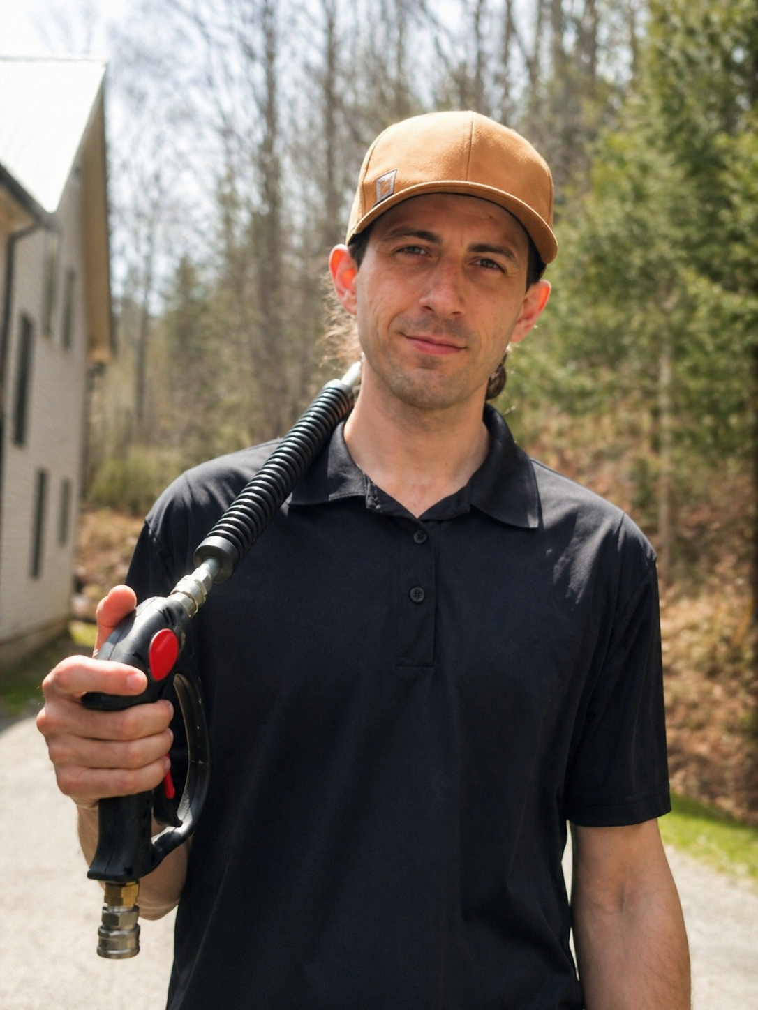 A man holding a pressure wash wand outdoors during daytime, wearing a brown cap and black shirt, with a background of trees and a house.