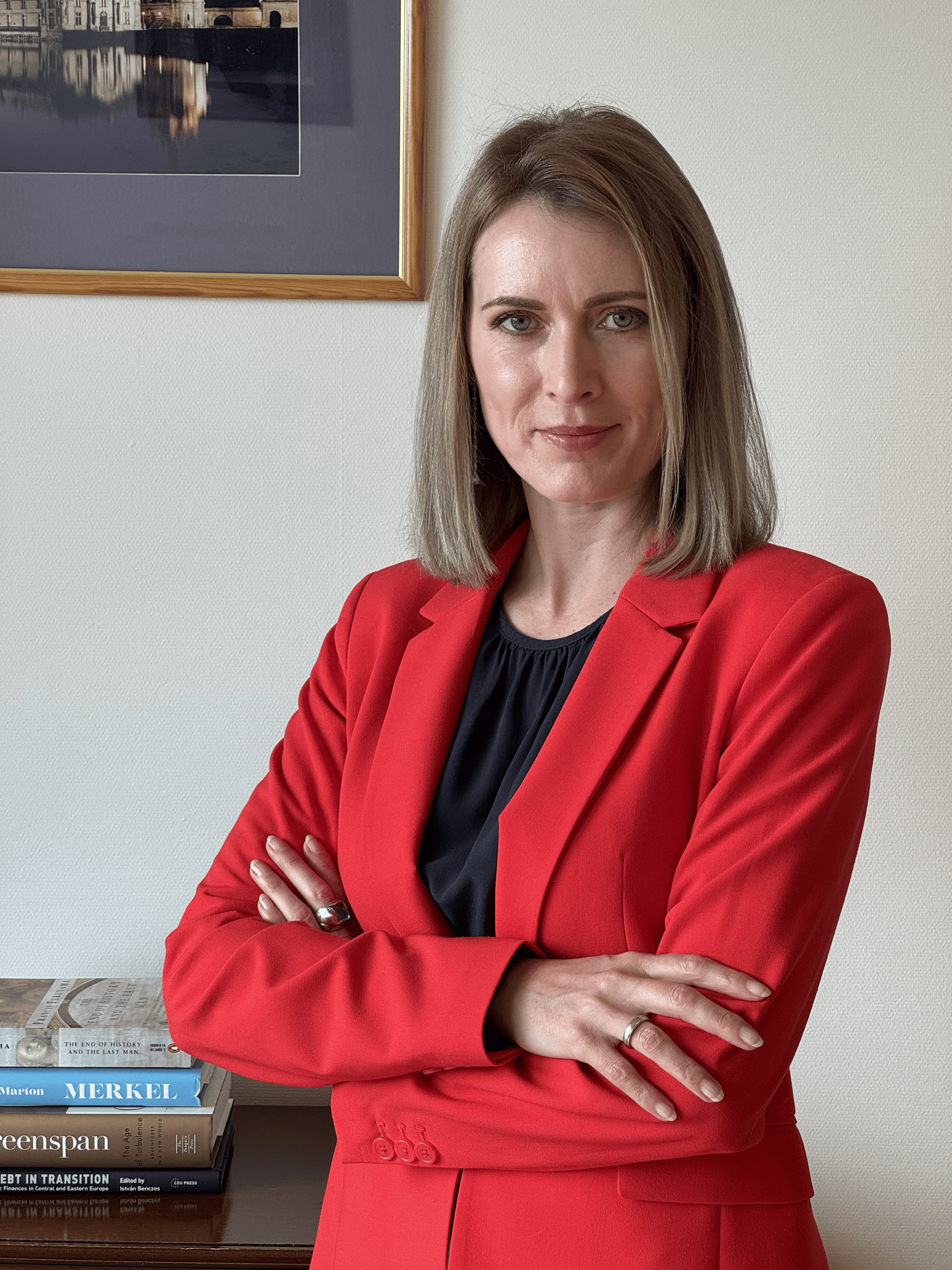 A woman in a red blazer standing with arms crossed in front of a white wall with framed artwork and a bookshelf.