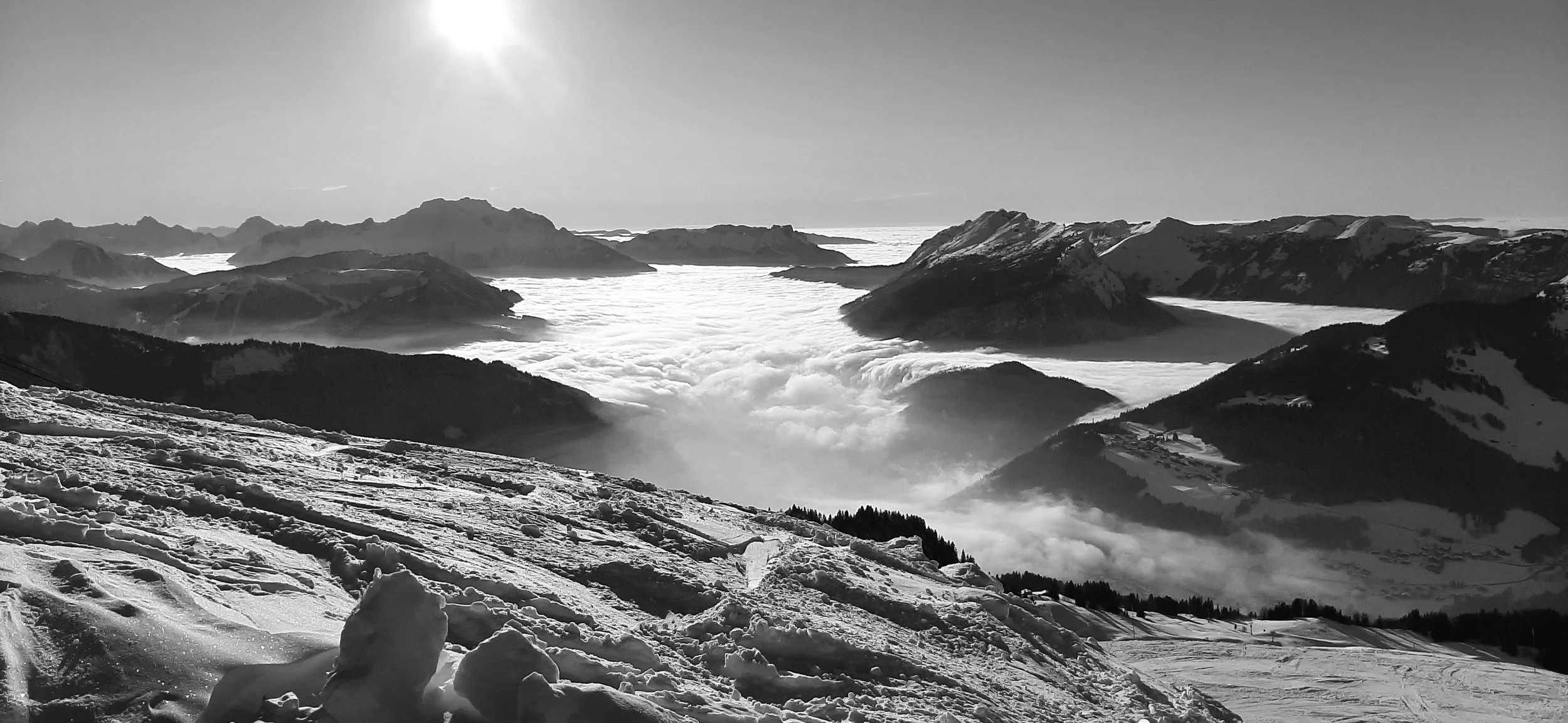 Snow-covered mountain landscape with peaks above a sea of clouds, sunny sky, and ski tracks in the foreground.