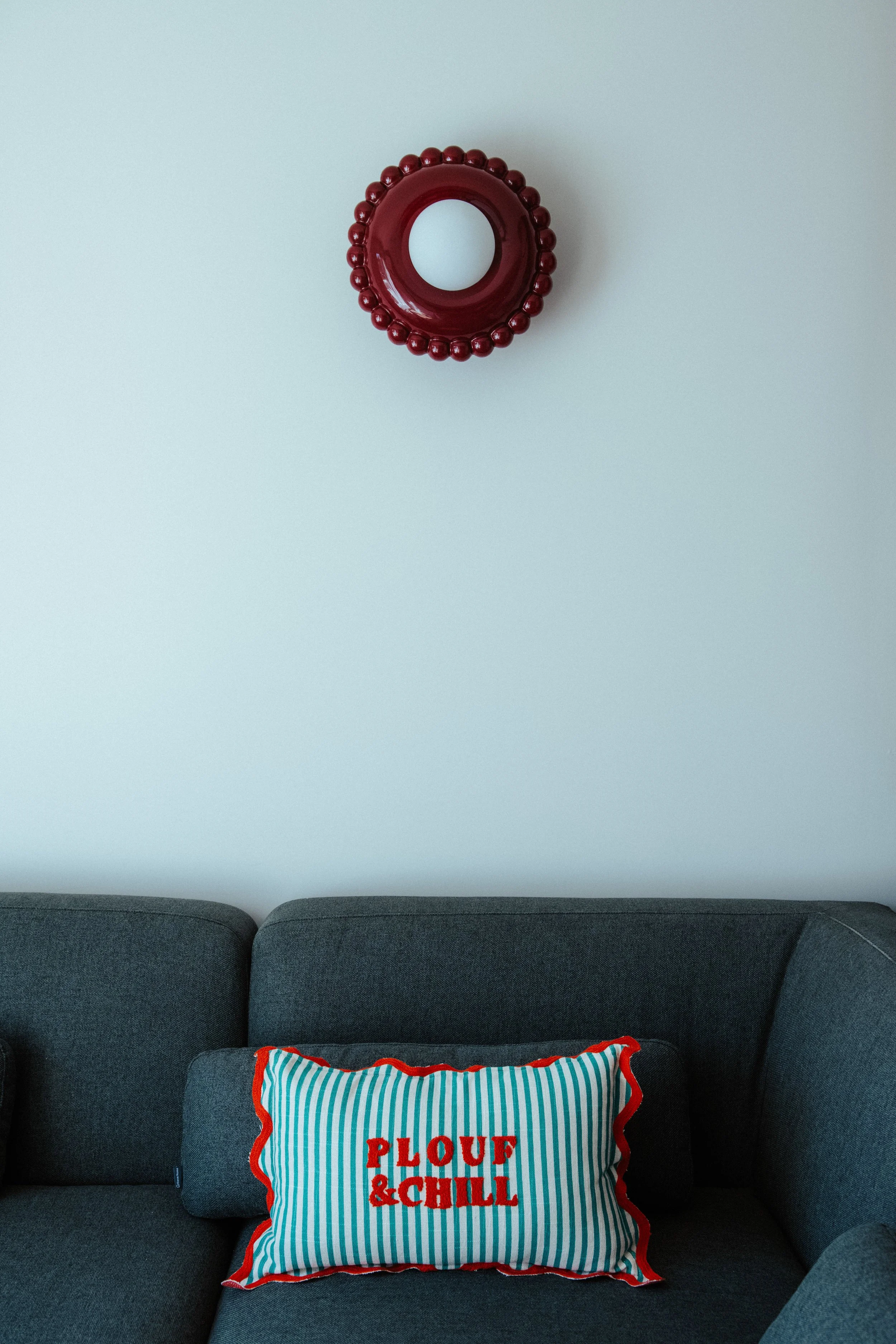 Coussin décoratif à rayures vertes et blanches avec le texte rouge 'PLOUF & CHILL' sur un canapé gris foncé, contre un mur blanc.