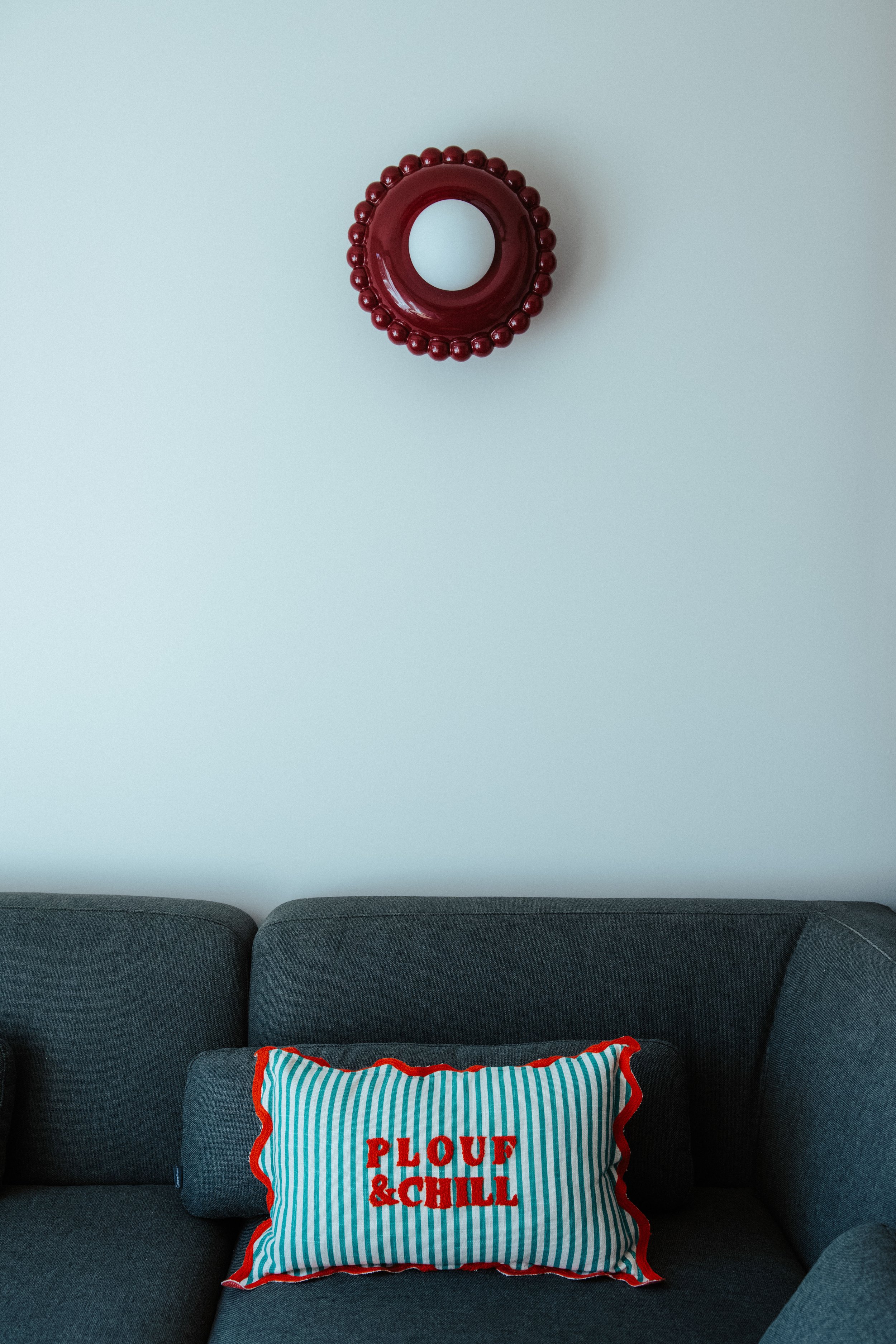 Un canapé gris foncé avec un coussin rayé bleu, blanc et rouge portant l'inscription 'PLOUF & CHILL', placé contre un mur blanc avec un miroir rond rouge entouré de boules rouges.