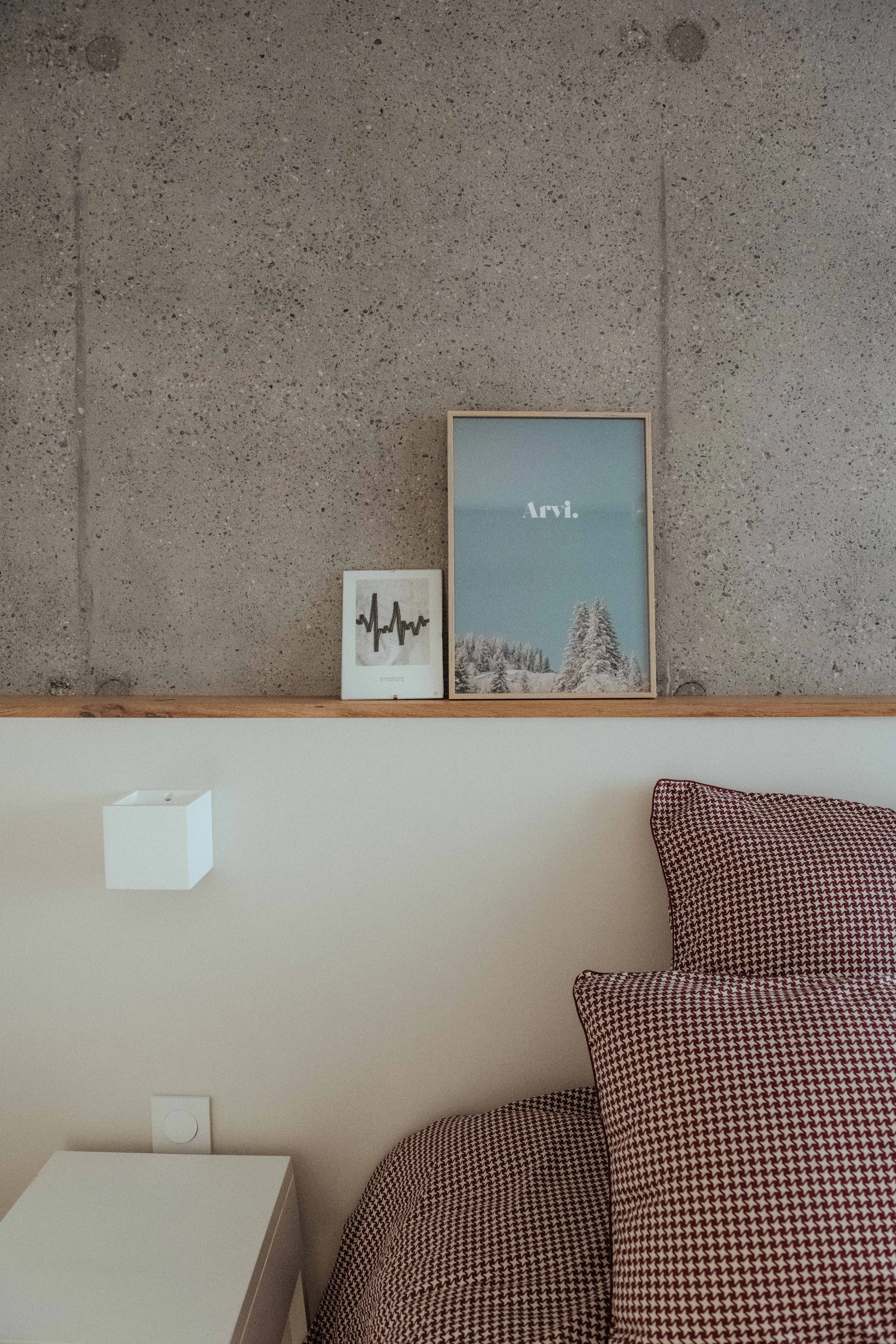 Une étagère en bois avec deux cadres photo et une lampe blanche au mur, une chaise avec un coussin à motif en zigzag rouge foncé et blanc.