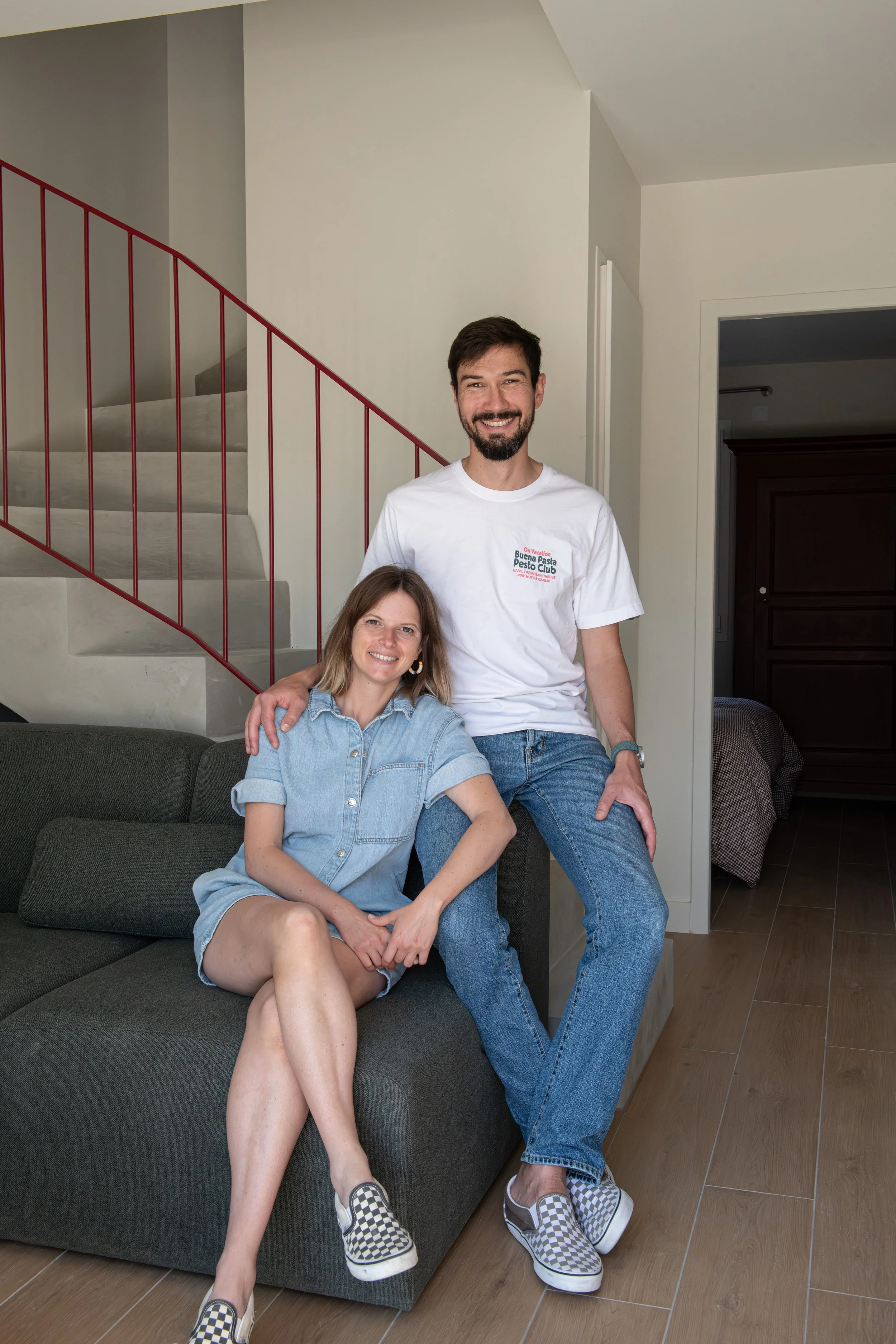 Un homme et une femme posent ensemble dans un intérieur moderne, la femme est assise sur un canapé et l'homme est debout derrière elle. La femme porte une robe en denim et des chaussures à motif écossais, l'homme porte un t-shirt blanc avec un texte et des chaussures à motif écossais. Le décor inclut une cage d'escalier en métal rouge et un coin de pièce avec un meuble en bois.