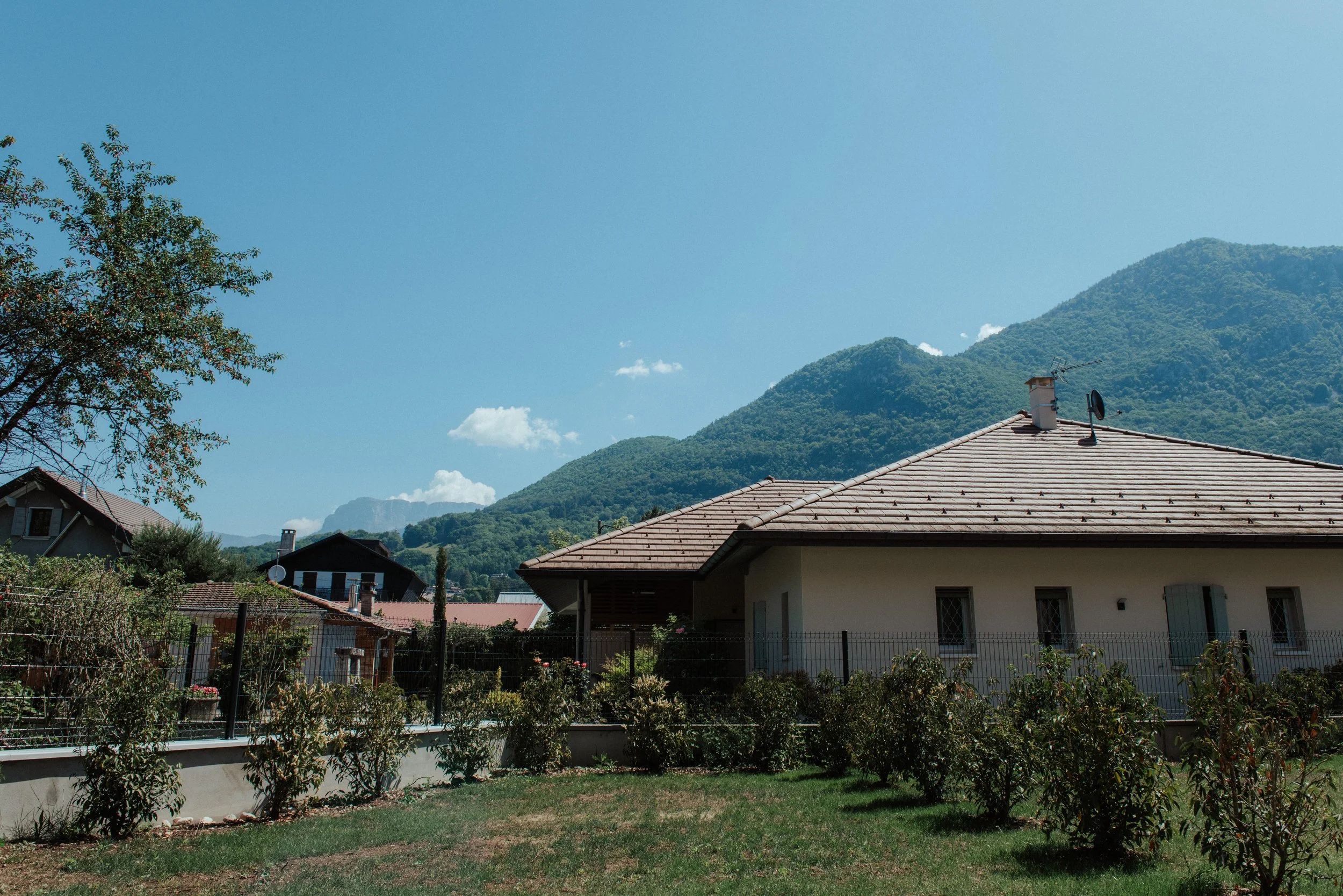 Une maison avec un toit en tuiles rouges surplombée par une cheminée, avec un jardin entouré d'une clôture et un arbre à gauche, avec des montagnes verdoyantes en arrière-plan sous un ciel bleu clair avec quelques nuages.