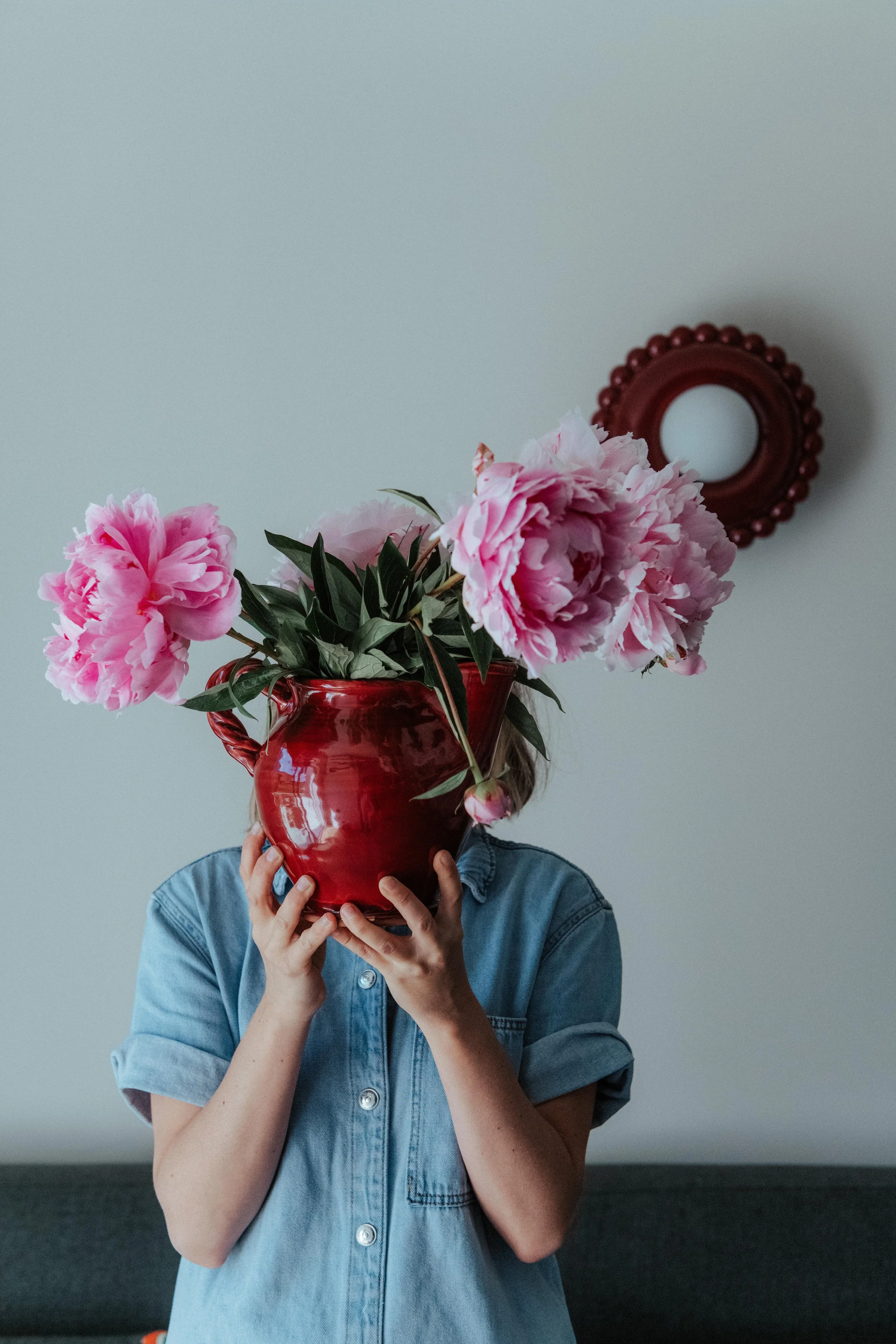 Une personne porte un bouquet de grandes fleurs roses dans un vase rouge, couvrant son visage, et se tient devant un mur blanc avec un miroir rond décoratif en arrière-plan.