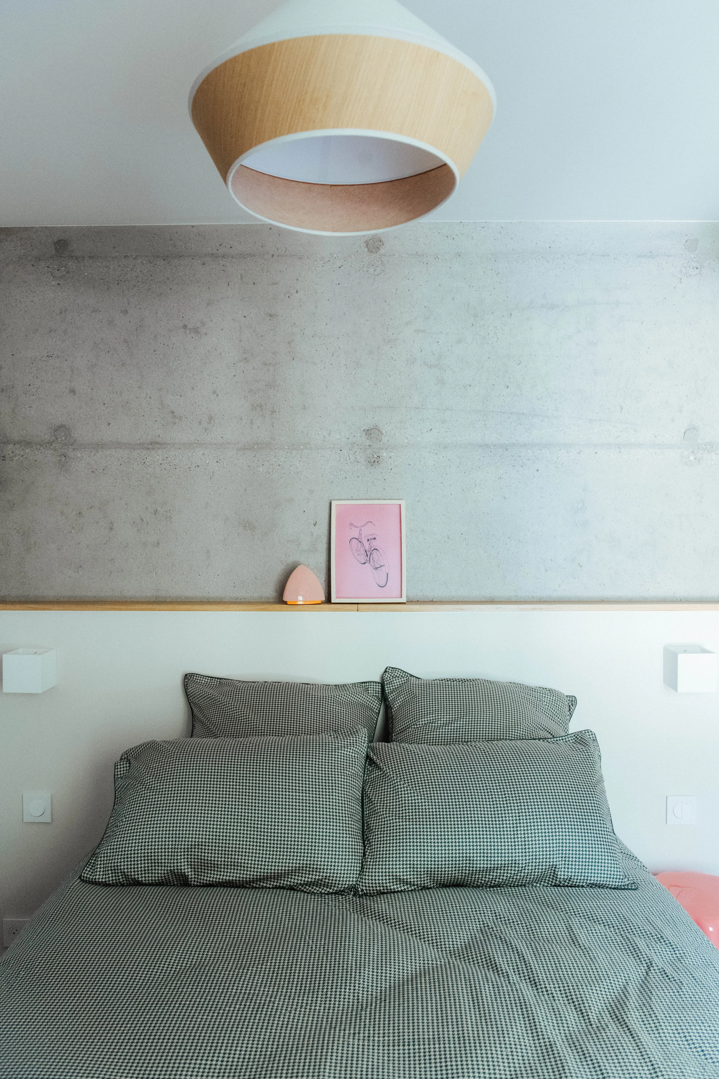 Un lit avec une couverture à motif pied-de-poule, deux oreillers assortis, deux appliques murales blanches, un portrait encadré avec un dessin de vélo, une lampe de chevet rose, dans une chambre moderne et épurée.