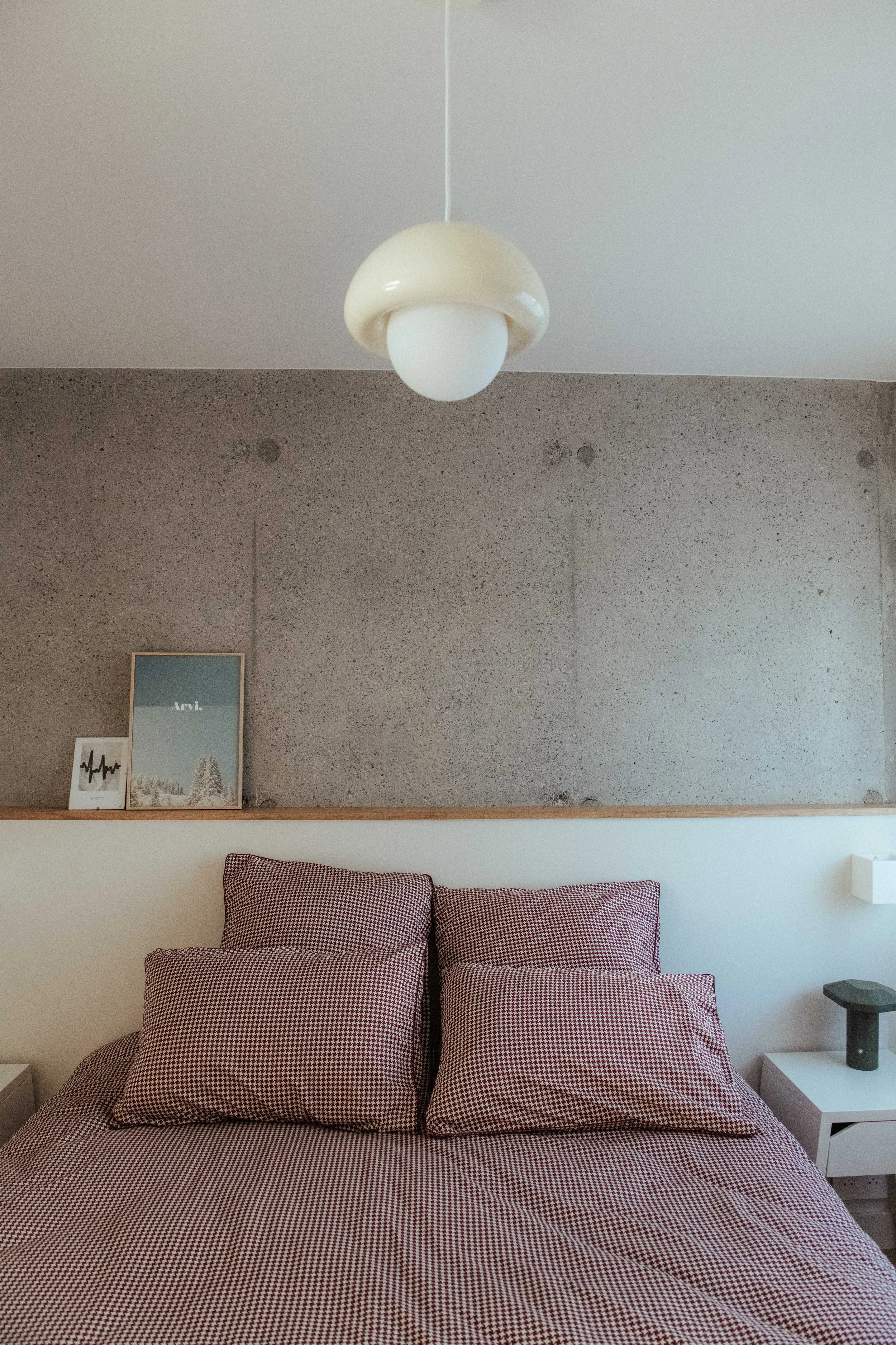 Un lit avec des draps et des coussins à motifs à carreaux rouges et blancs, dans une chambre moderne avec une décoration minimaliste, une table de chevet blanche, un cadre photo, et une lampe