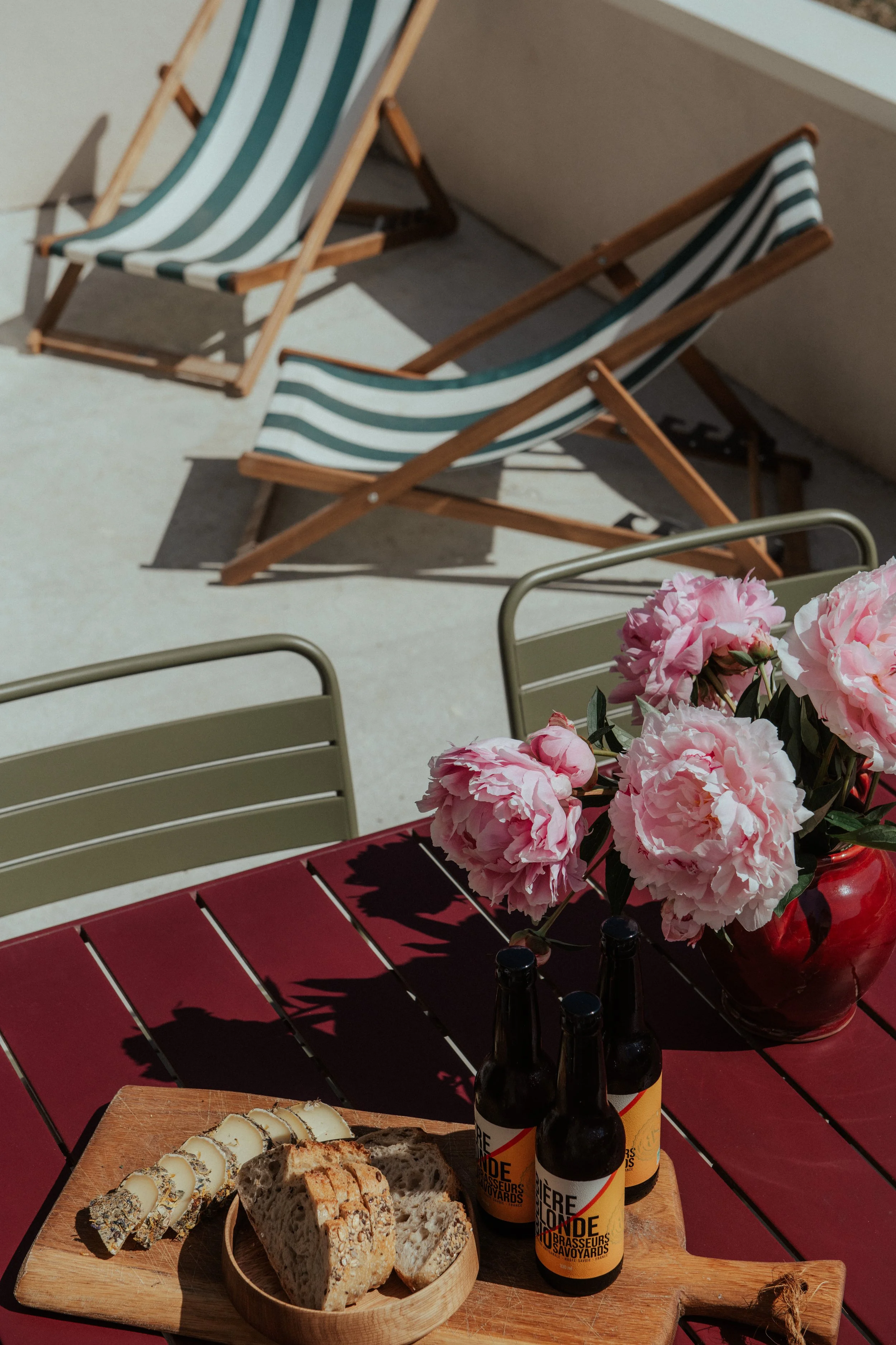 Balcon avec chaises en bois, table rouge avec un bouquet de fleurs roses, bouteilles de bière, plateau avec pain et fromage, en plein soleil.