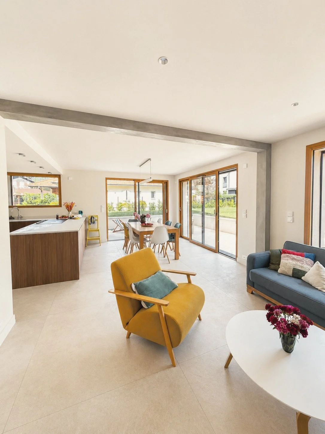 Intérieur moderne d'une maison avec salon, salle à manger lumineuse, grandes fenêtres, canapé bleu, fauteuil jaune et cuisine en arrière-plan.