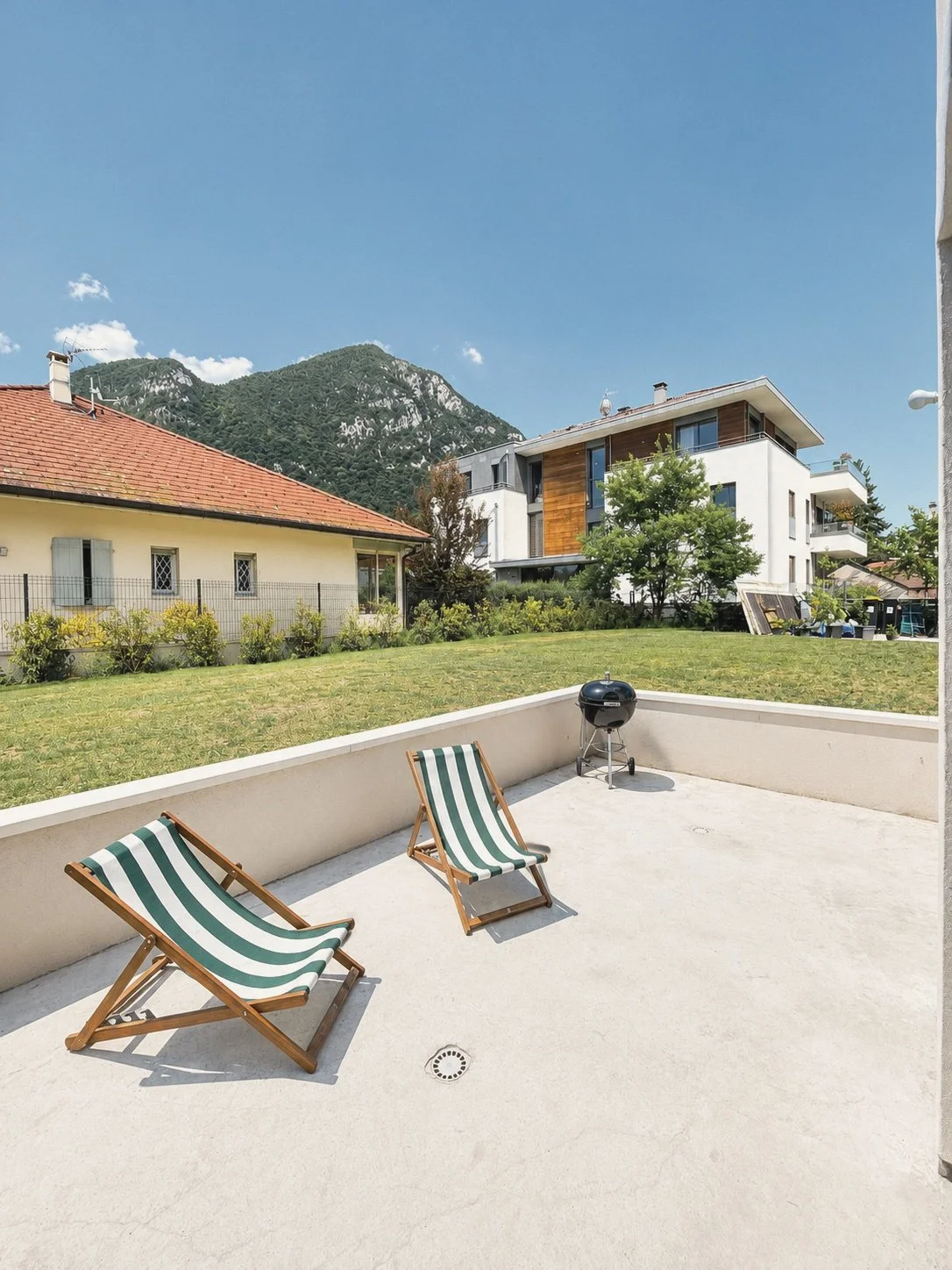 Terrasse ensoleillée avec deux chaises longues à rayures vertes et blanches, barbecue noir, pelouse verte, maisons modernes et montagnes en arrière-plan.