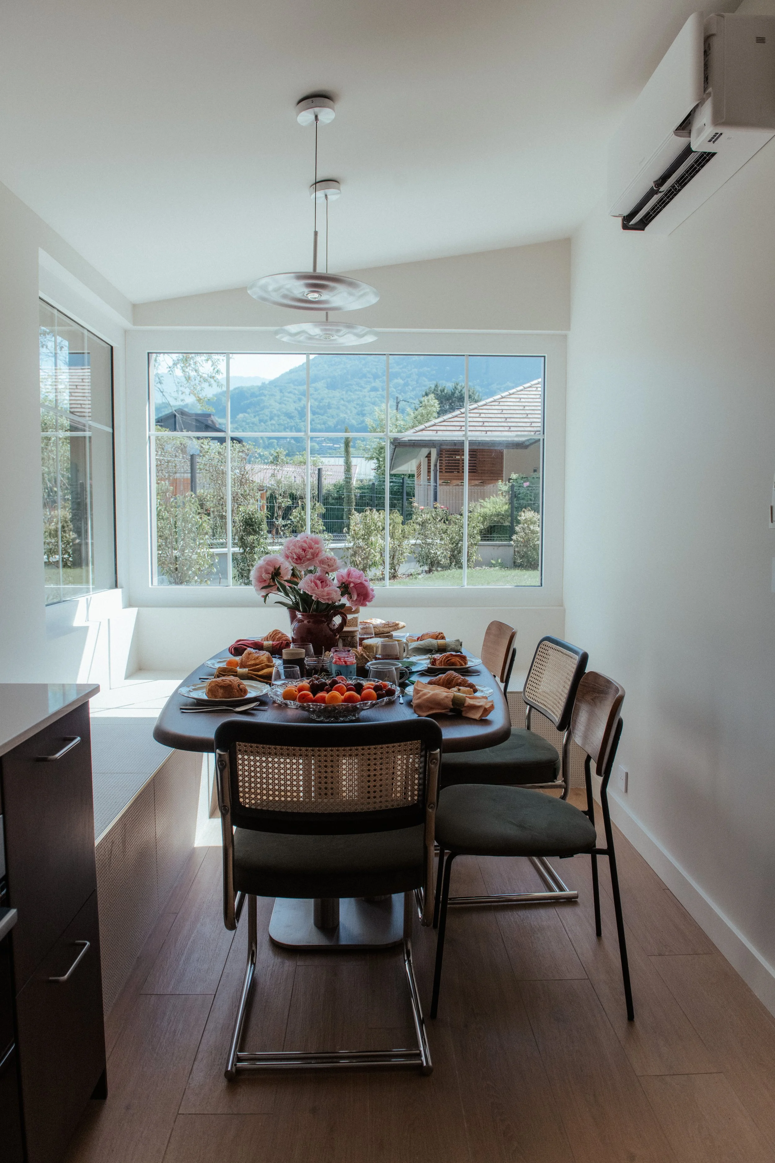 Une salle à manger éclairée par la lumière naturelle d'une grande fenêtre, avec une table dressée pour le petit déjeuner. La table comporte une vase avec des fleurs roses, des plats avec des croissants, des fruits, et des boissons. Il y a cinq chaises autour de la table, certaines avec assise en tissu gris et d'autres en bois avec assise en canevas.