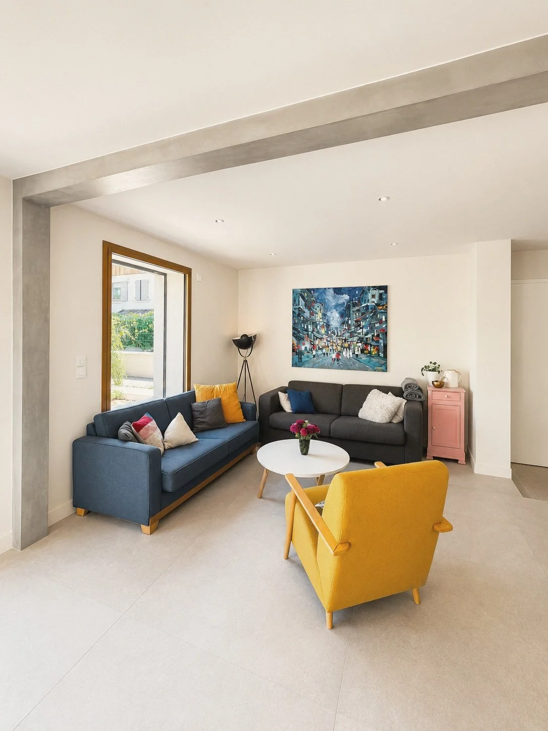 Salon moderne avec deux canapés gris, un fauteuil jaune, une table ronde blanche avec un vase de fleurs, une peinture colorée sur le mur, et une grande fenêtre laissant entrer la lumière naturelle.