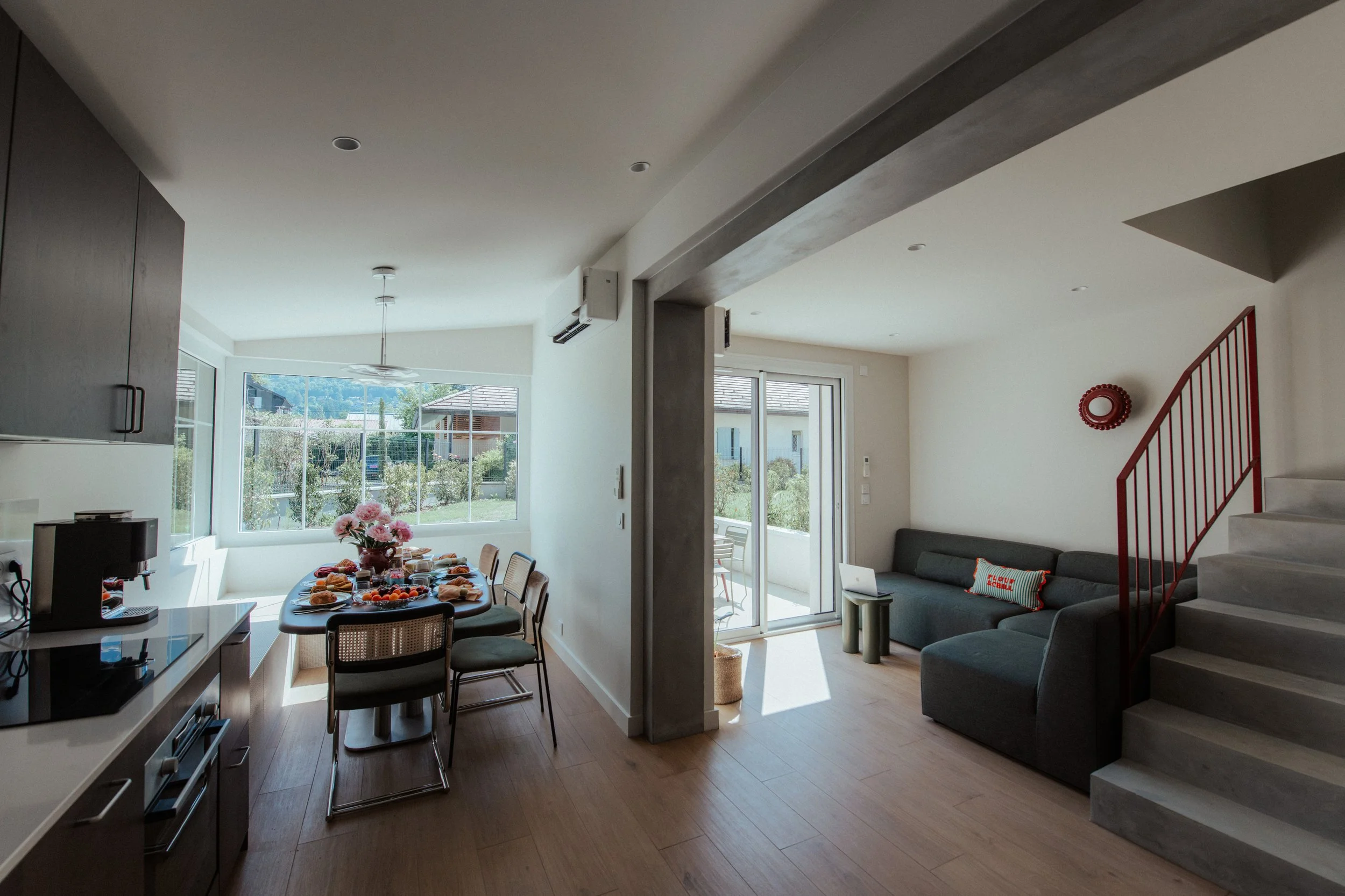 Intérieur d'une maison moderne avec salon et salle à manger, grandes fenêtres laissant entrer la lumière naturelle, décoration minimaliste, escalier en béton, canapé gris, table avec petit-déjeuner, vue sur le jardin extérieur.
