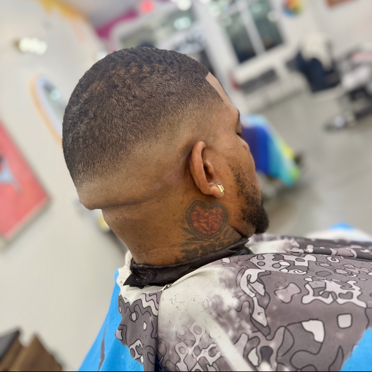 A man with short textured hair, a facial tattoo on his neck, and a diamond earring, sitting in a barber shop, possibly getting a haircut.