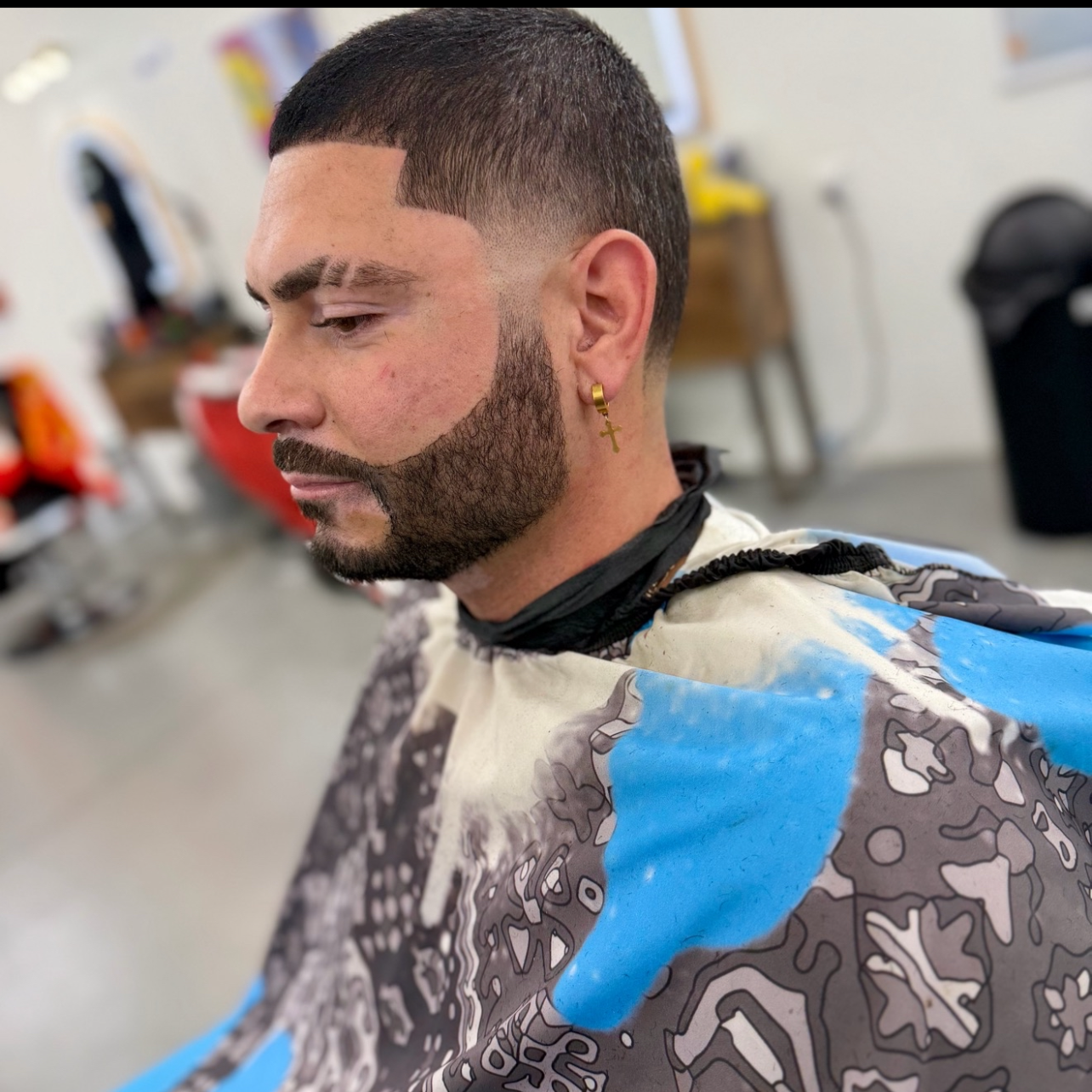 Man in a barber shop with a new hairstyle and beard, wearing a barber cape with a colorful abstract pattern, gold cross earring, and a black neck strap.