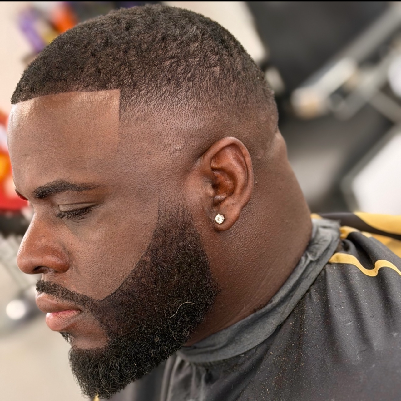 A man with a precise fade haircut and a full beard getting a haircut at a barbershop. He is wearing a black barber cape with yellow trim and has an earring in his left ear.