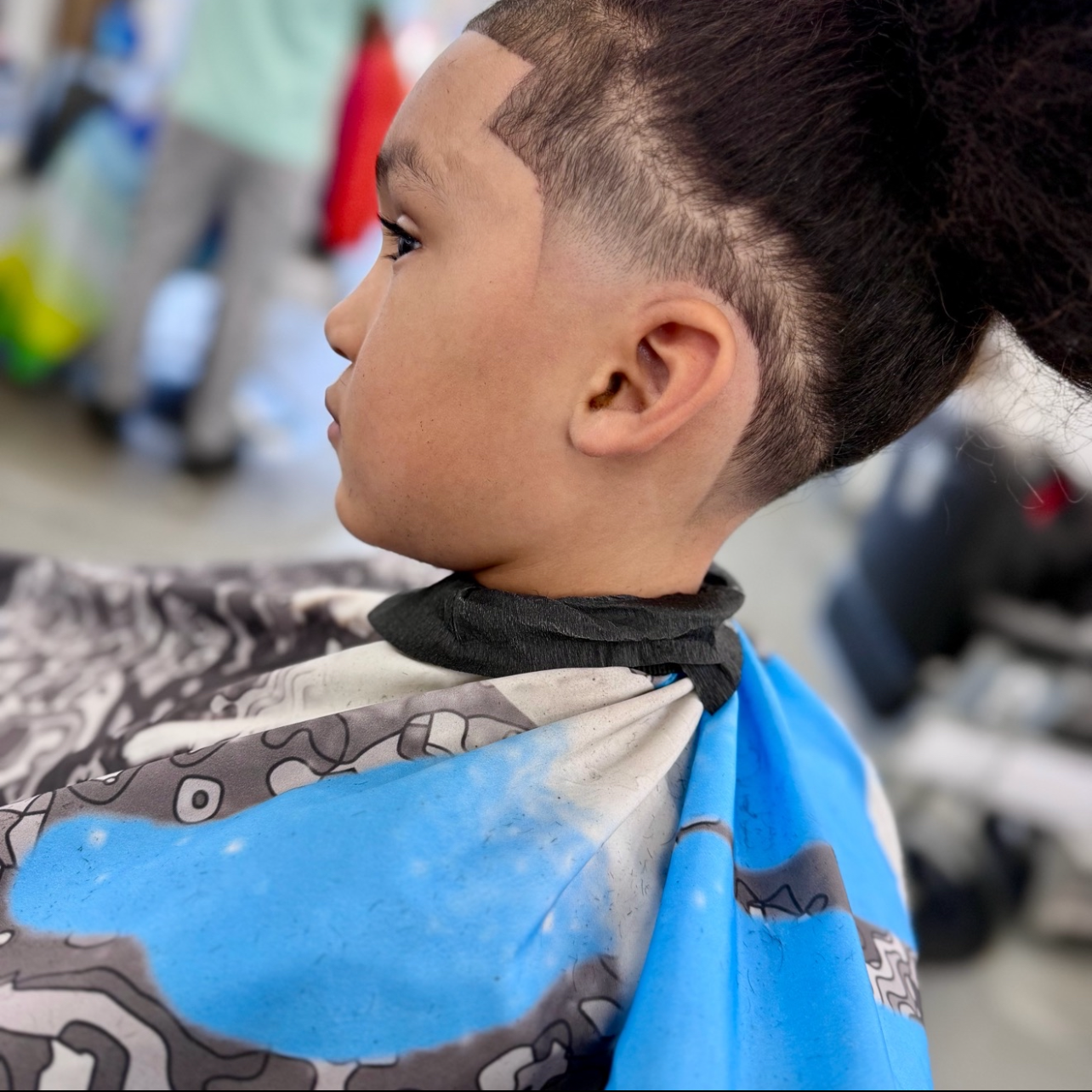 Young person sitting in a barber's chair with a fresh haircut, wearing a cape with a pattern, inside a barber shop.