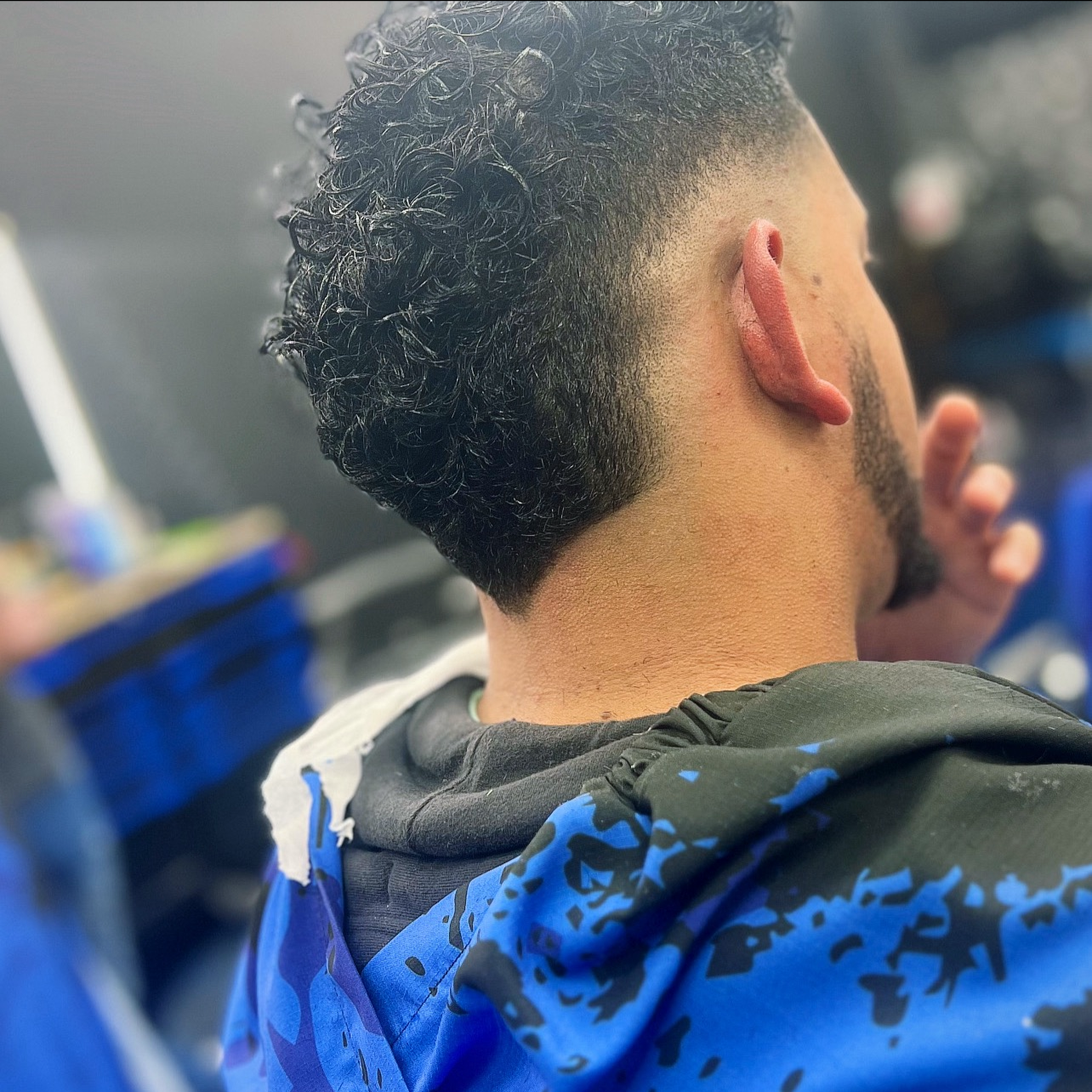 A person with curly hair and a fade haircut, wearing a blue and black hoodie, sitting in what appears to be a barbershop.