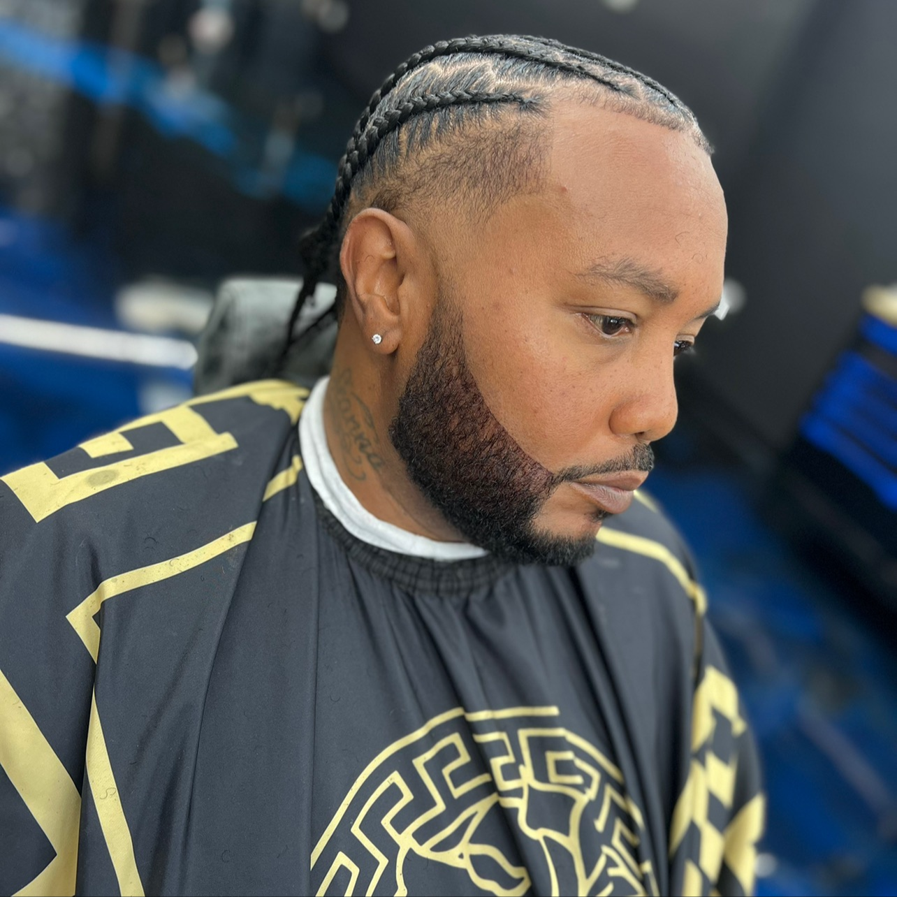 A man with braided hairstyle and a beard, wearing a black and gold patterned cape, sitting in a barbershop chair.