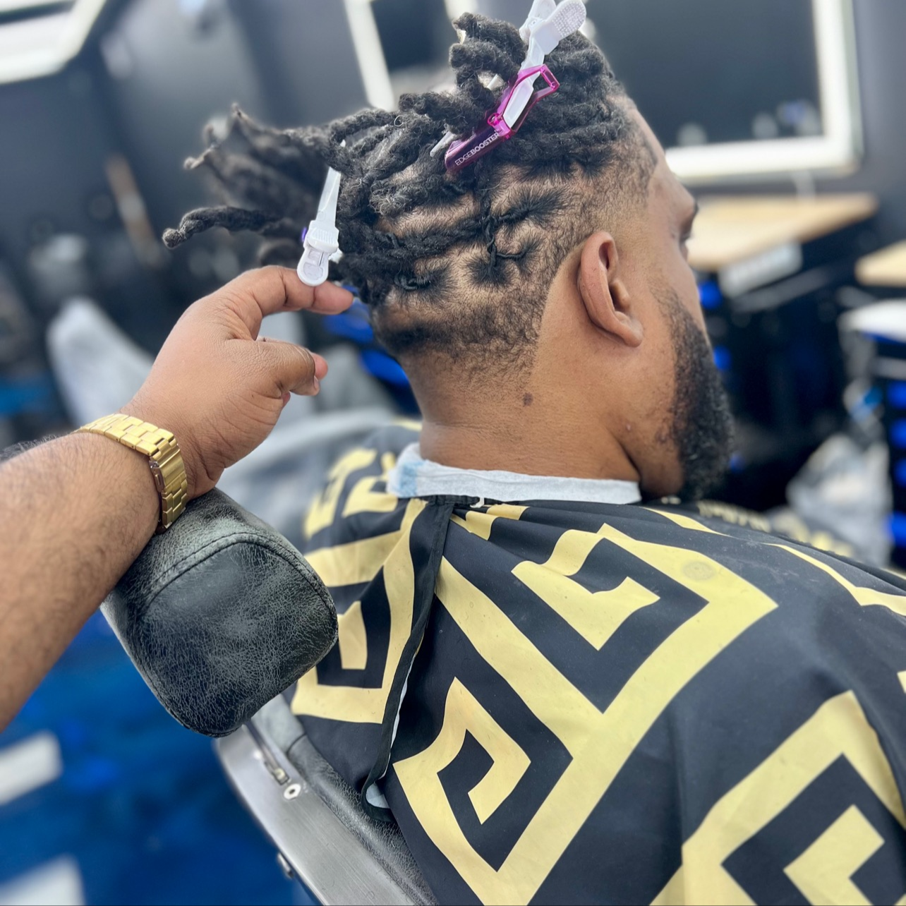 A man with dreadlocks getting a haircut at a barbershop, wearing a black and yellow patterned cape.