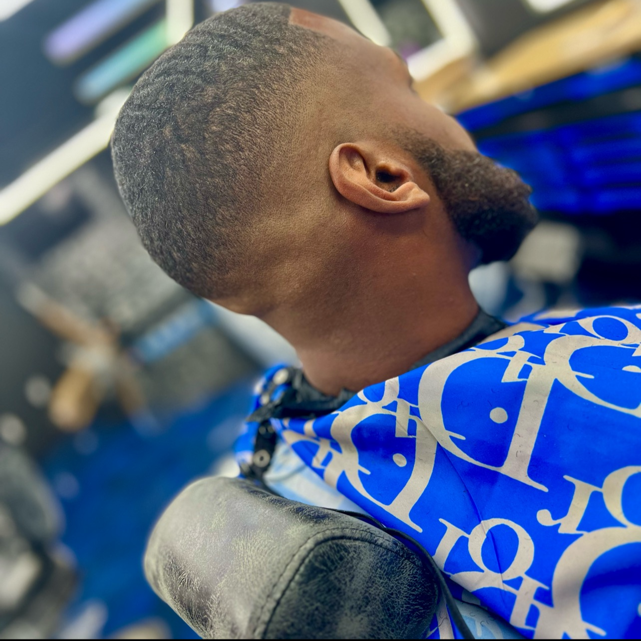 A man sitting in a barber chair, with a fresh haircut, wearing a blue and white barber cape with abstract patterns.