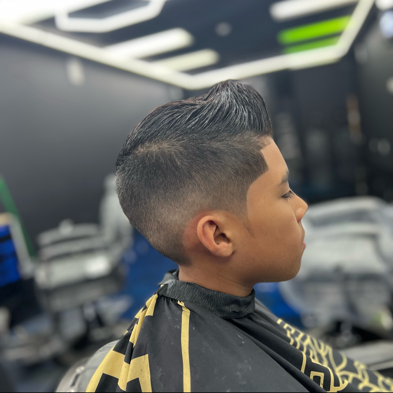 Young man with a modern tapered haircut, sitting in a barbershop chair with a black and yellow cape, getting a haircut.