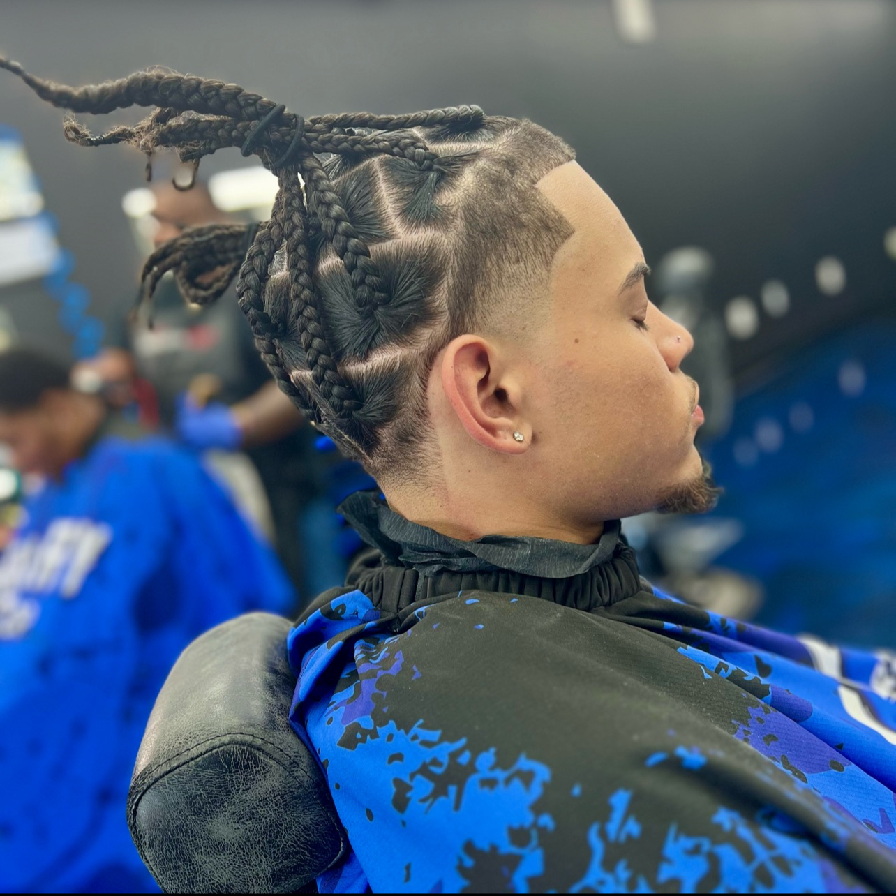 Person with braided hair and shaved sides relaxing with eyes closed in a barbershop or salon.