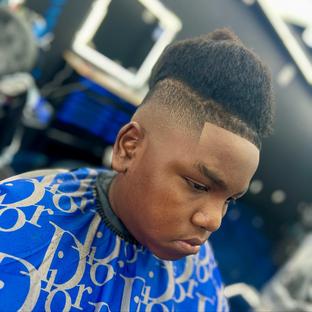 Close-up of a young man with a fresh, high fade haircut, looking down, wearing a blue barber cape with white lettering, in a barbershop setting.