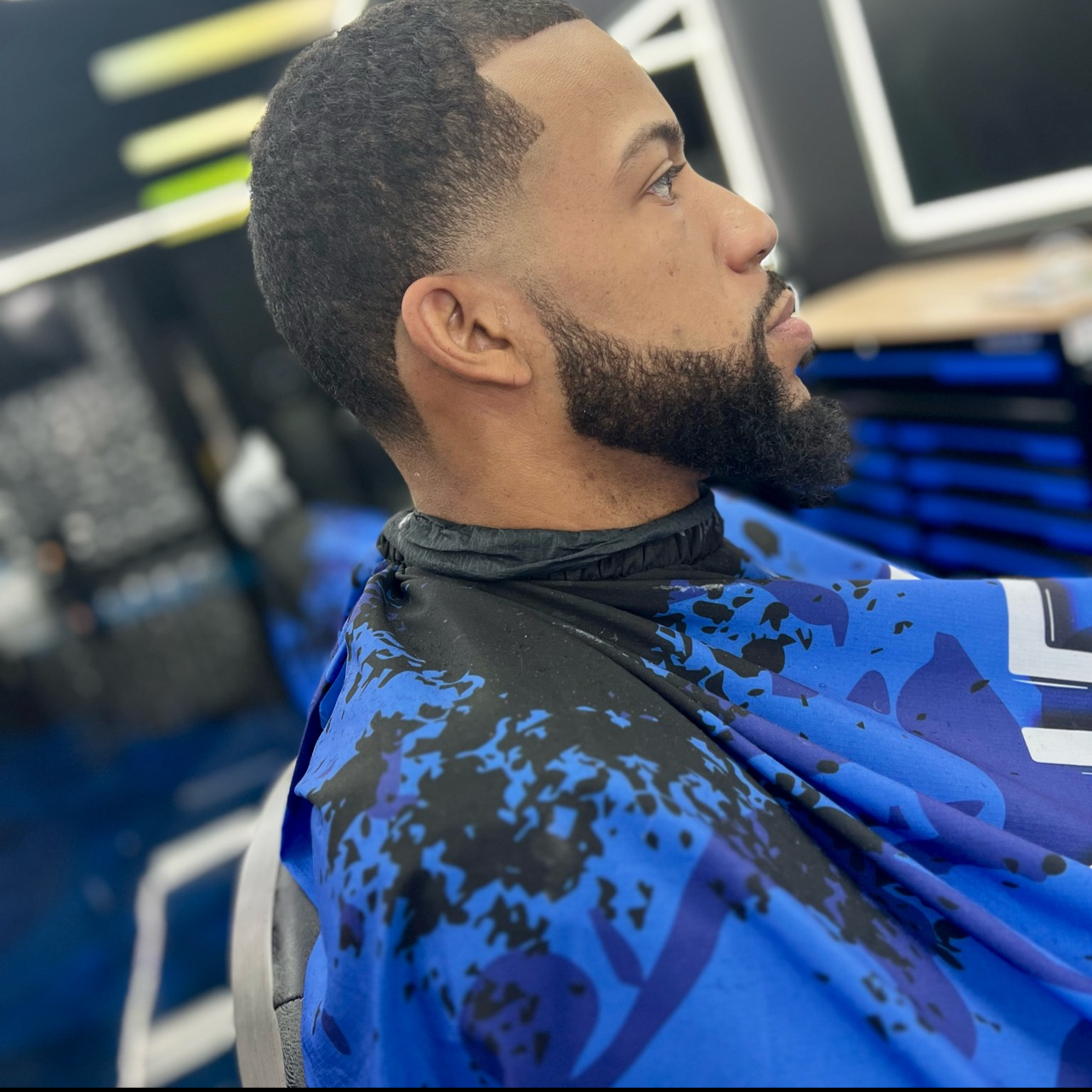 A man with a beard and short curly hair sitting in a barber's chair wearing a blue and black barber cape, likely getting a haircut at a barbershop.