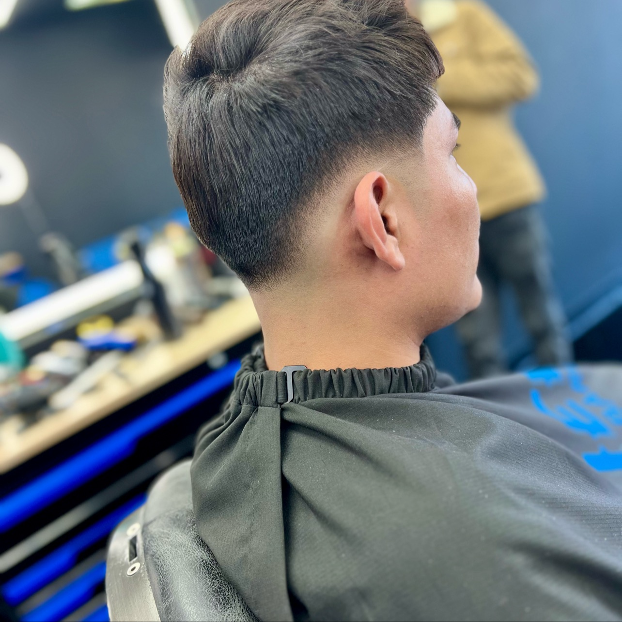Side view of a young man with a fresh, short hairstyle in a barbershop. He is seated in a barber's chair, wearing a black cape, with a blurred background of barber tools and a mirror.