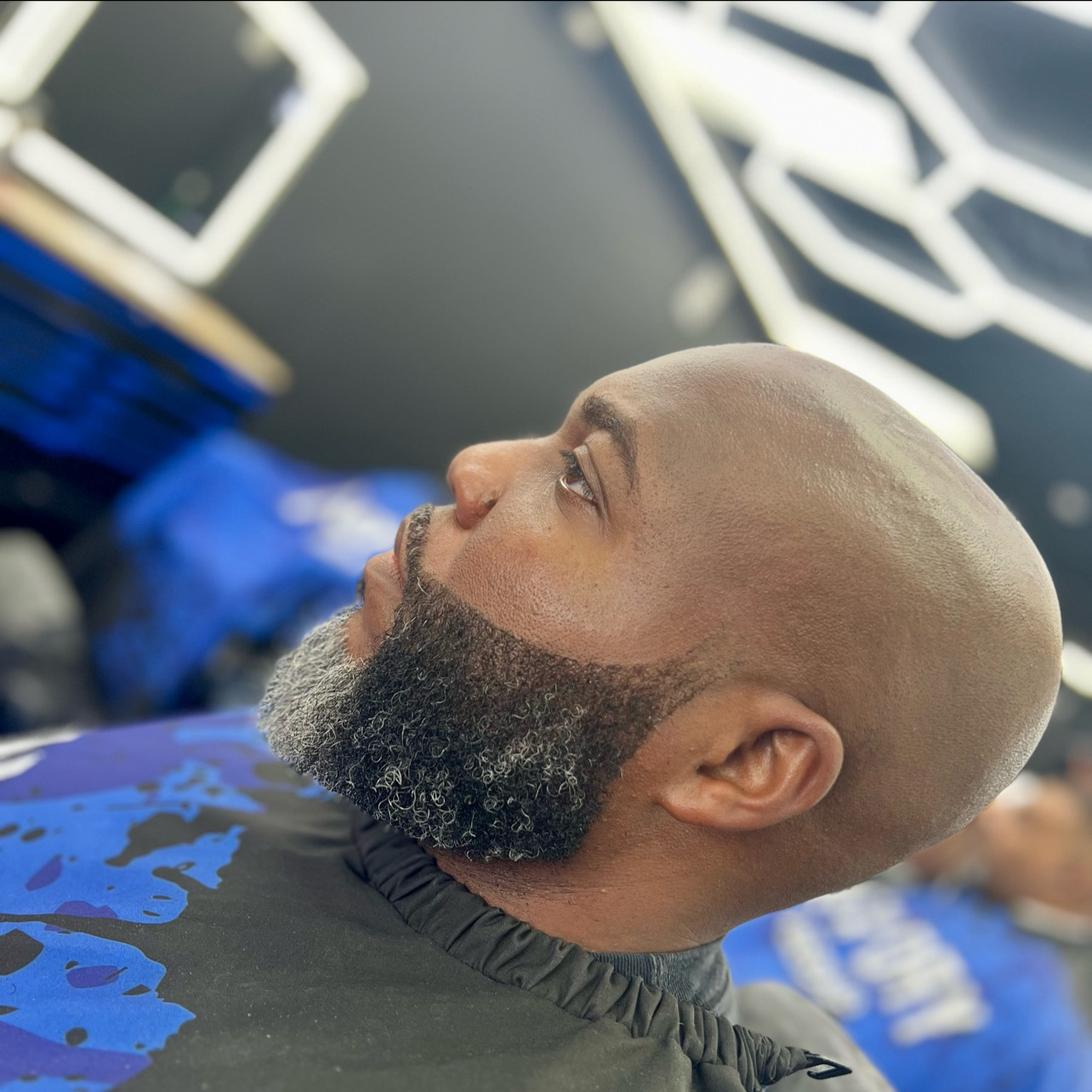 A man with a bald head and a beard looking upward while lying in a barbershop chair, with hair-cutting tools and a barber's gloves visible in the background.