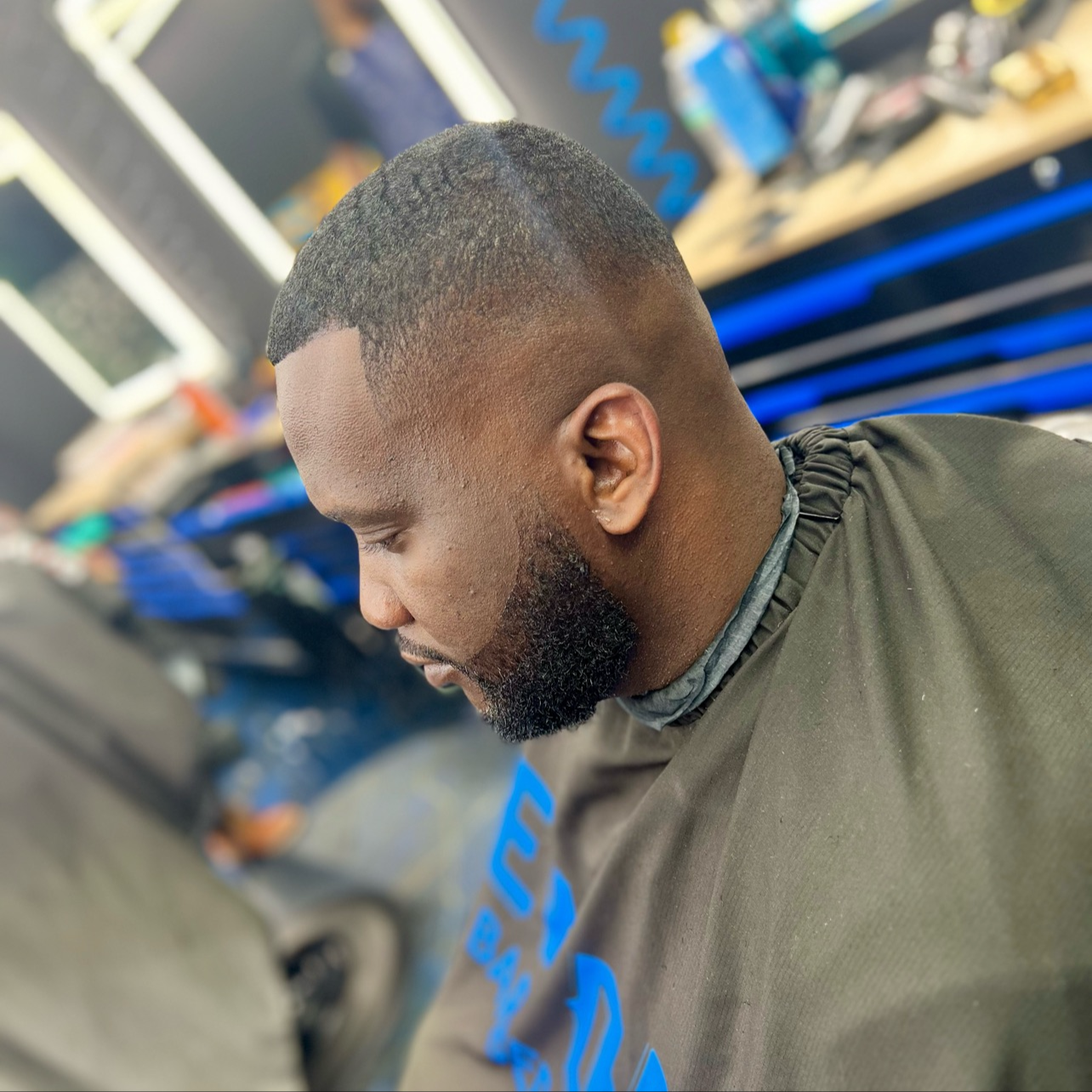 A young man with a fresh haircut, a beard, and a dark hair fade, is seated in a barbershop with barber equipment and products visible in the background.