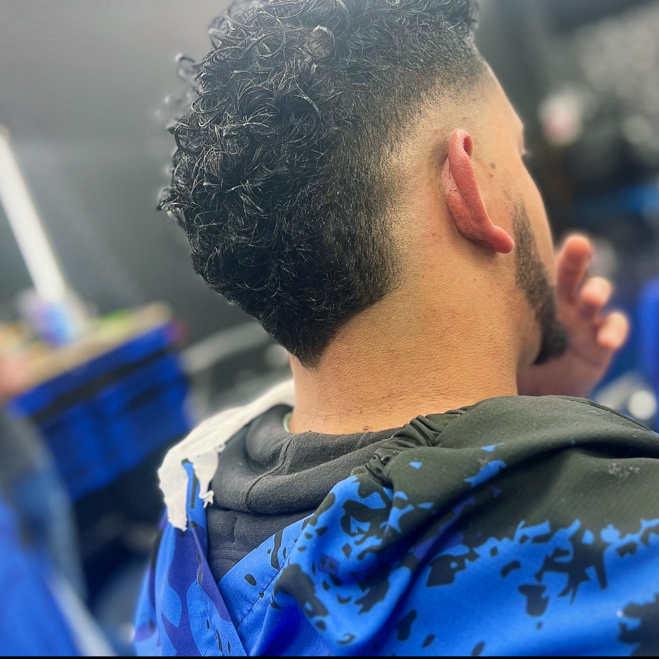 A person with short curly black hair and tan skin, sitting in a barber shop. The person is wearing a black and blue patterned hoodie and has a large novelty tongue earrings on the ear visible. Blurred background with blue and black objects.