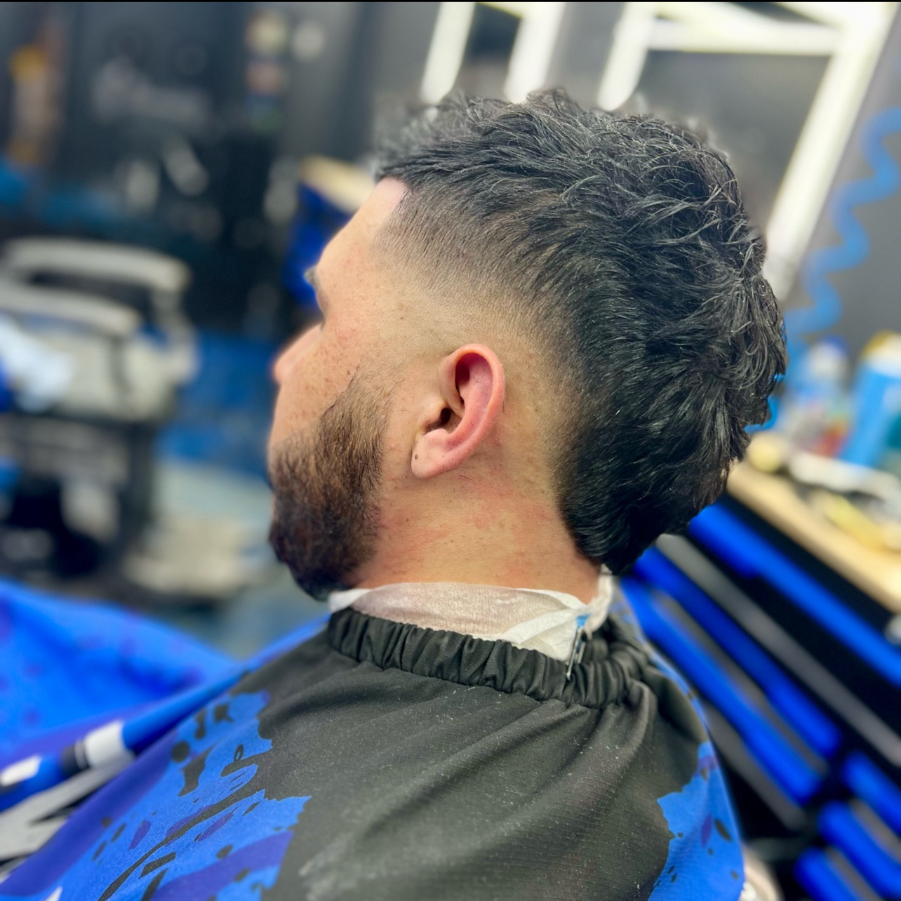 Side view of a man with a fresh high fade haircut, styled with curly hair on top and faded sides, sitting in a barbershop.