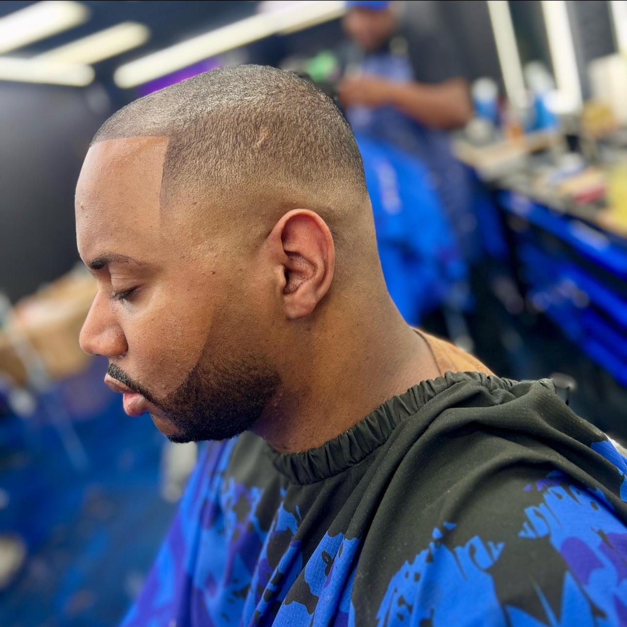 Side profile of a man with a freshly shaved haircut, sitting in a barbershop. The background shows blurred items of barber tools and another person in a blue apron.