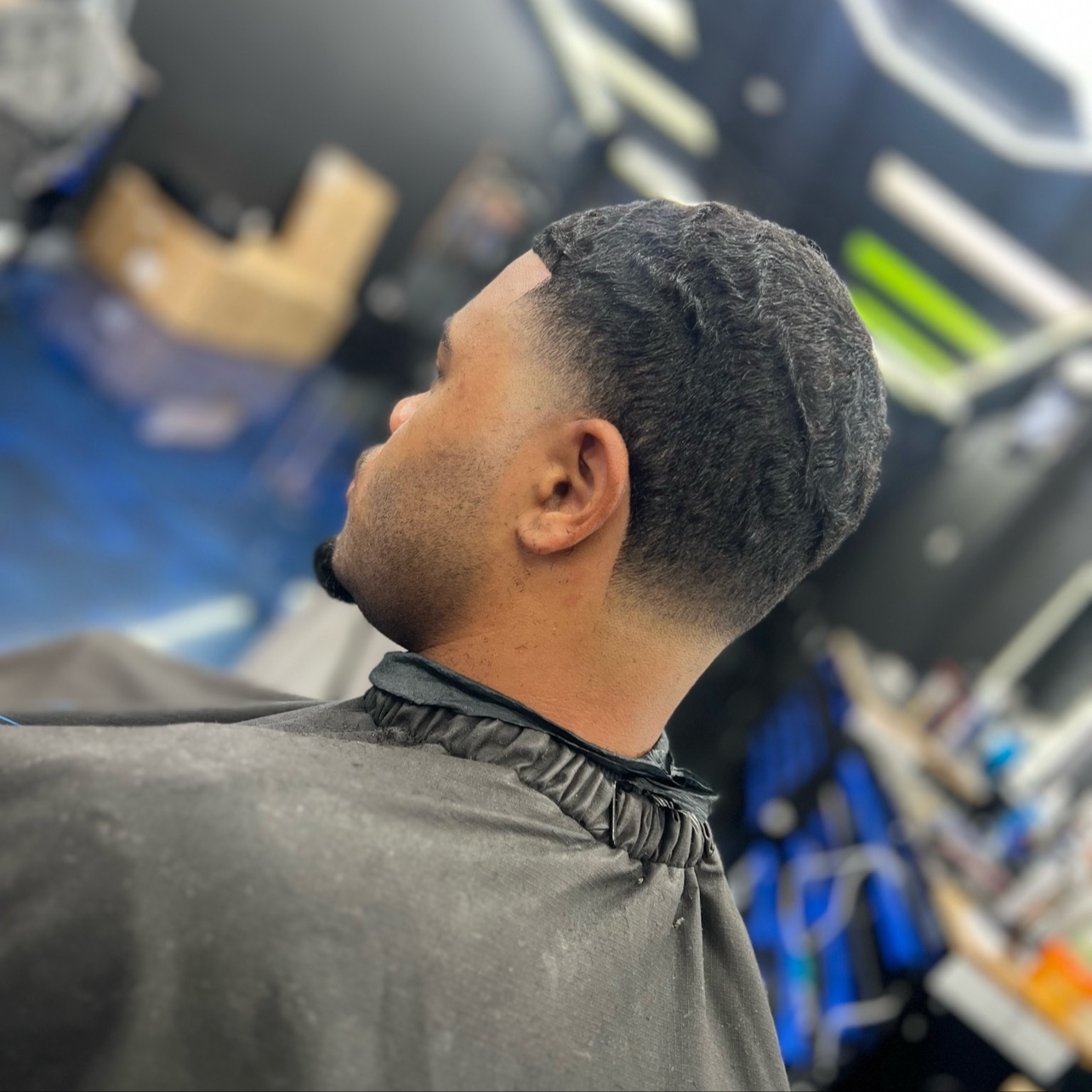 Man with a textured, wavy haircut in a barbershop, wearing a black barber cape, with blurred barber tools and products in the background.