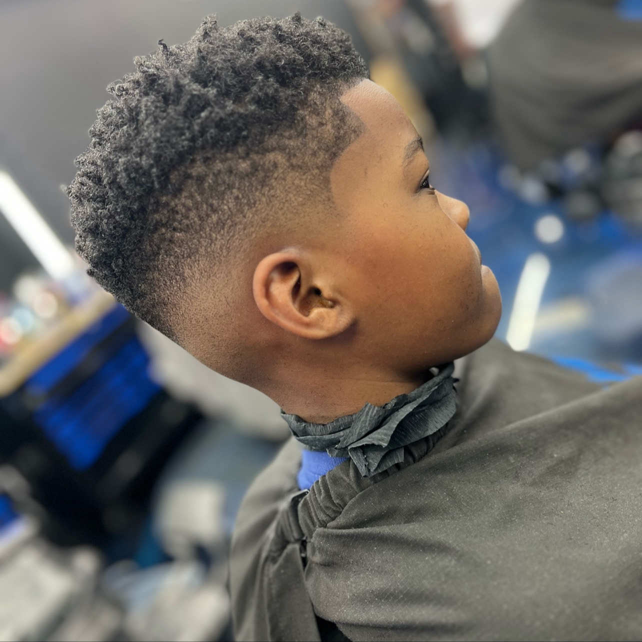 A young boy with a textured, natural hairstyle sitting in a barbershop chair, looking to the side.