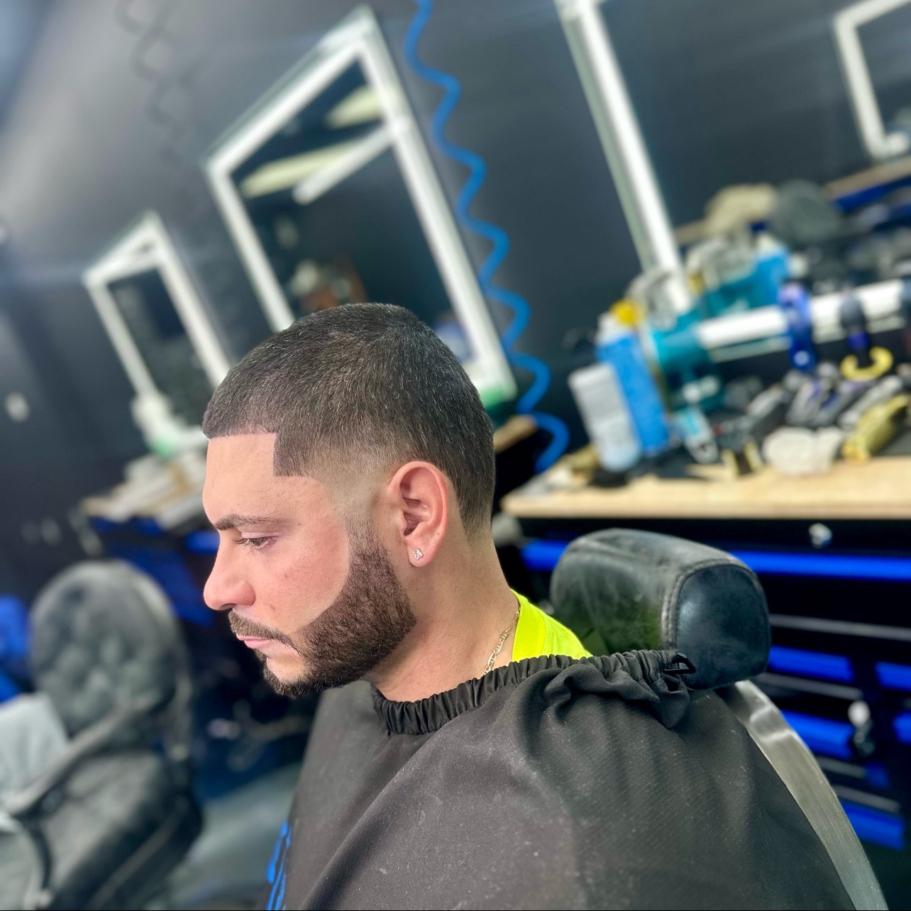 A man with a fresh fade haircut and beard sits in a barber chair in a barbershop, looking down with a serious expression, with various barber tools and supplies visible in the background.