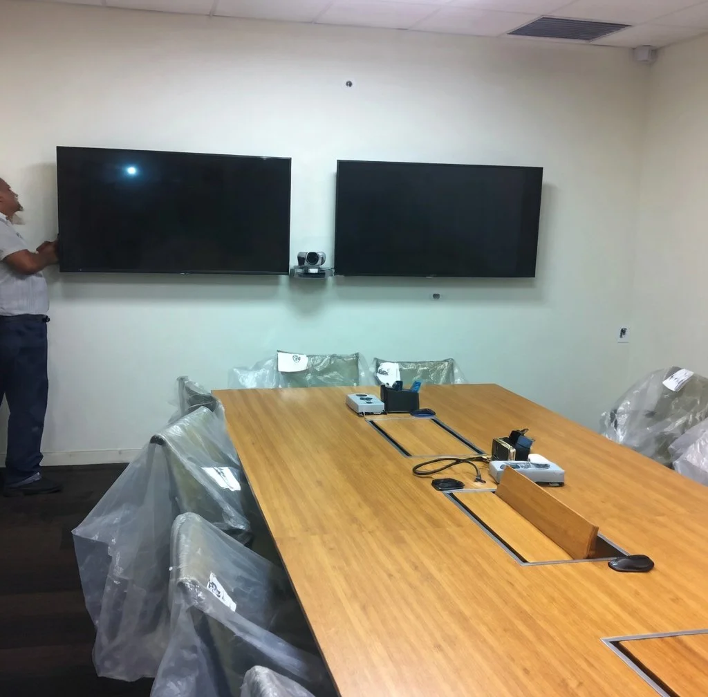 A conference room with a long wooden table, six chairs covered in plastic, two large black wall-mounted televisions, and a man standing to the left.
