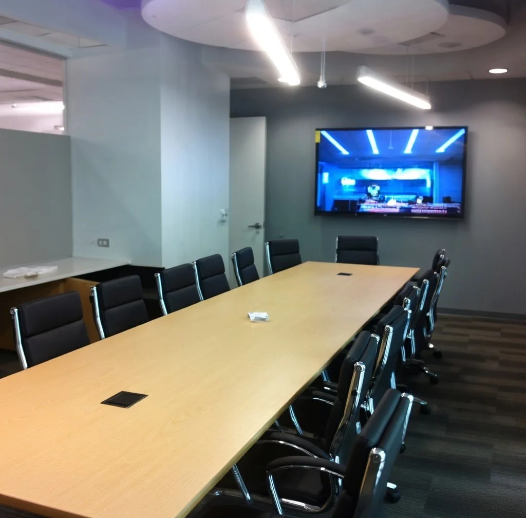 Empty conference room with a long wooden table, black leather chairs, a wall-mounted TV displaying a blue-lit room, and modern lighting in the ceiling.