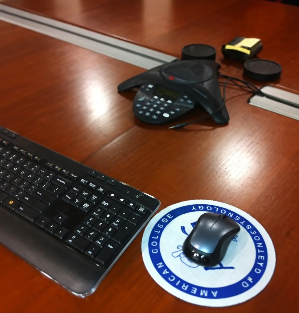 Office desk with a black keyboard, wireless mouse on a round blue and white sticker, conference phone, and other office items on a wooden surface.