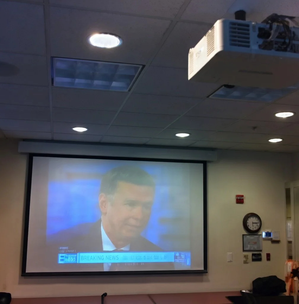 A projection screen inside a room showing a man in a suit during a news broadcast. A ceiling-mounted projector is visible in the upper right corner, and a clock, fire alarm, and control panel are on the wall to the right of the screen.