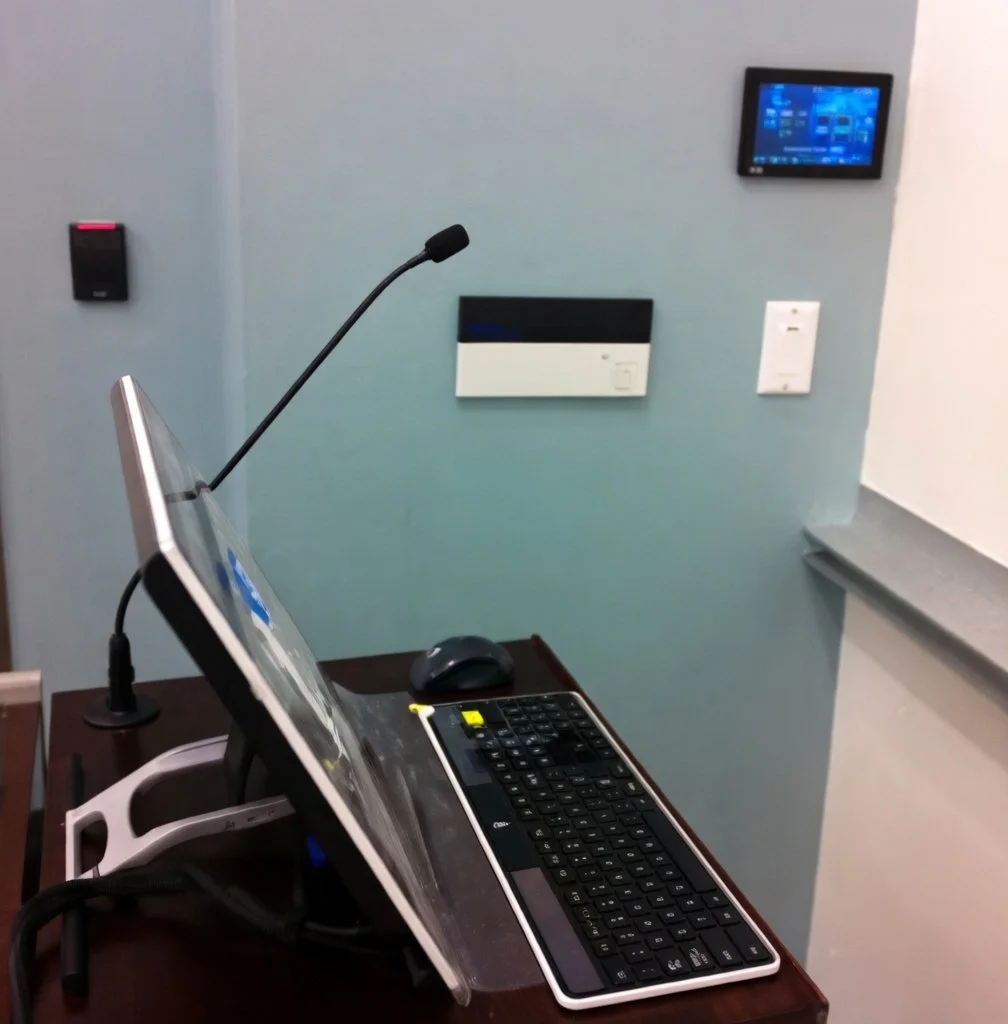 A medical or office desk setup with a touchscreen computer monitor, keyboard, mouse, and microphone, located in front of a wall with various electronic devices and controls.