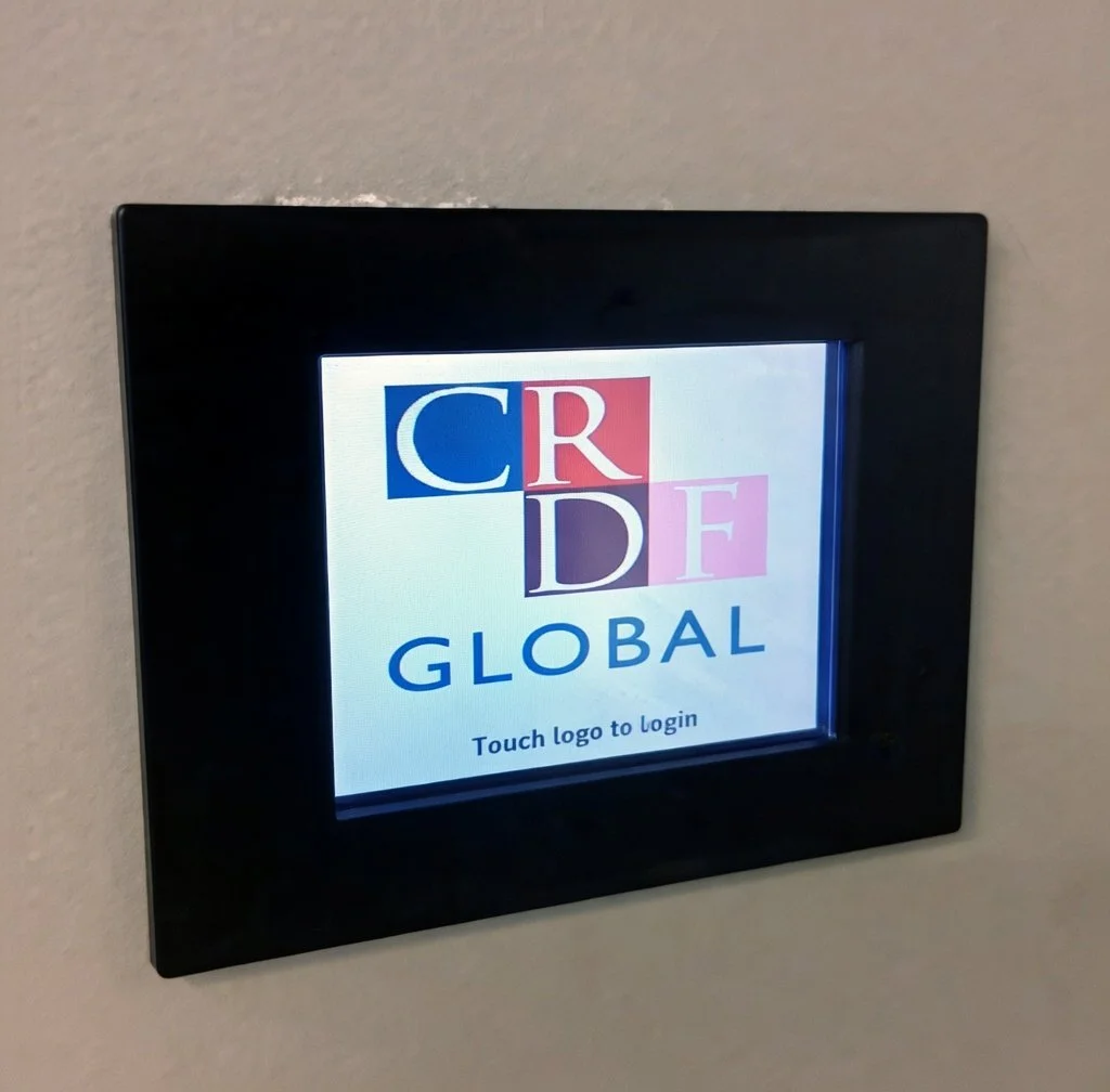 Digital touchscreen with a logo that reads 'C R D F GLOBAL' and the instruction 'Touch logo to login' on a beige wall.