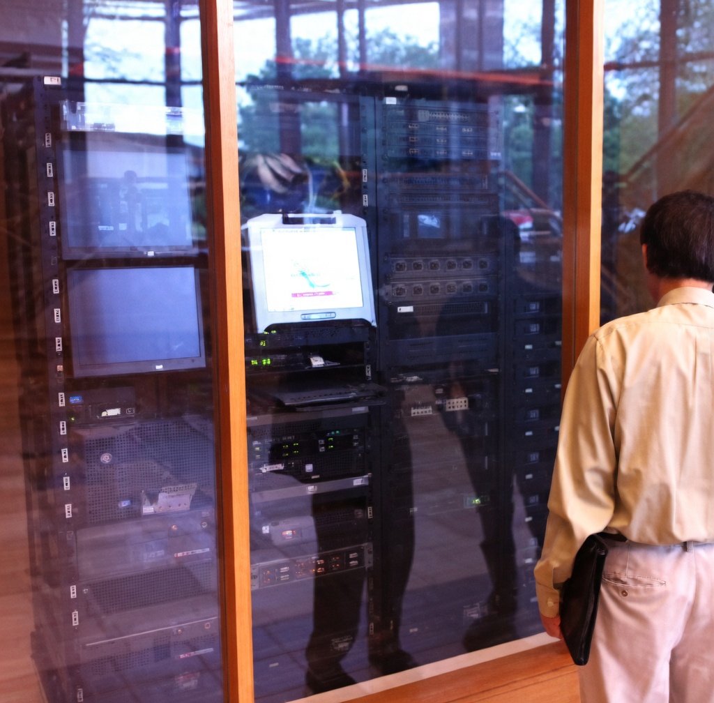 A person standing outside a glass enclosure containing server racks and IT equipment, with reflections and trees visible through the glass.