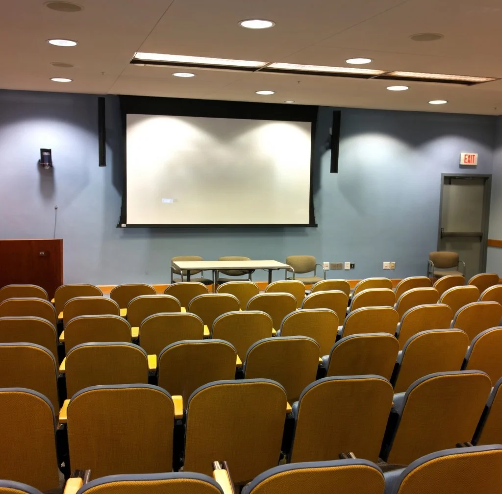 An empty lecture hall with rows of yellow chairs, a large blank white screen on the front wall, and a small table with two chairs facing the audience. There is an exit door on the right side of the room.
