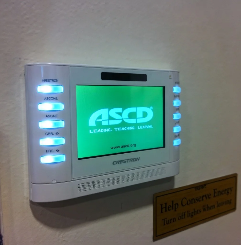 An electronic display panel mounted on a wall with blue indicator lights and the ASCD logo on the screen; a yellow energy conservation sign is partially visible below it.