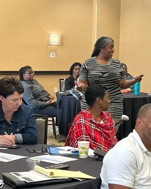 A group of people sitting at a conference table in a meeting room, with one woman standing and speaking.