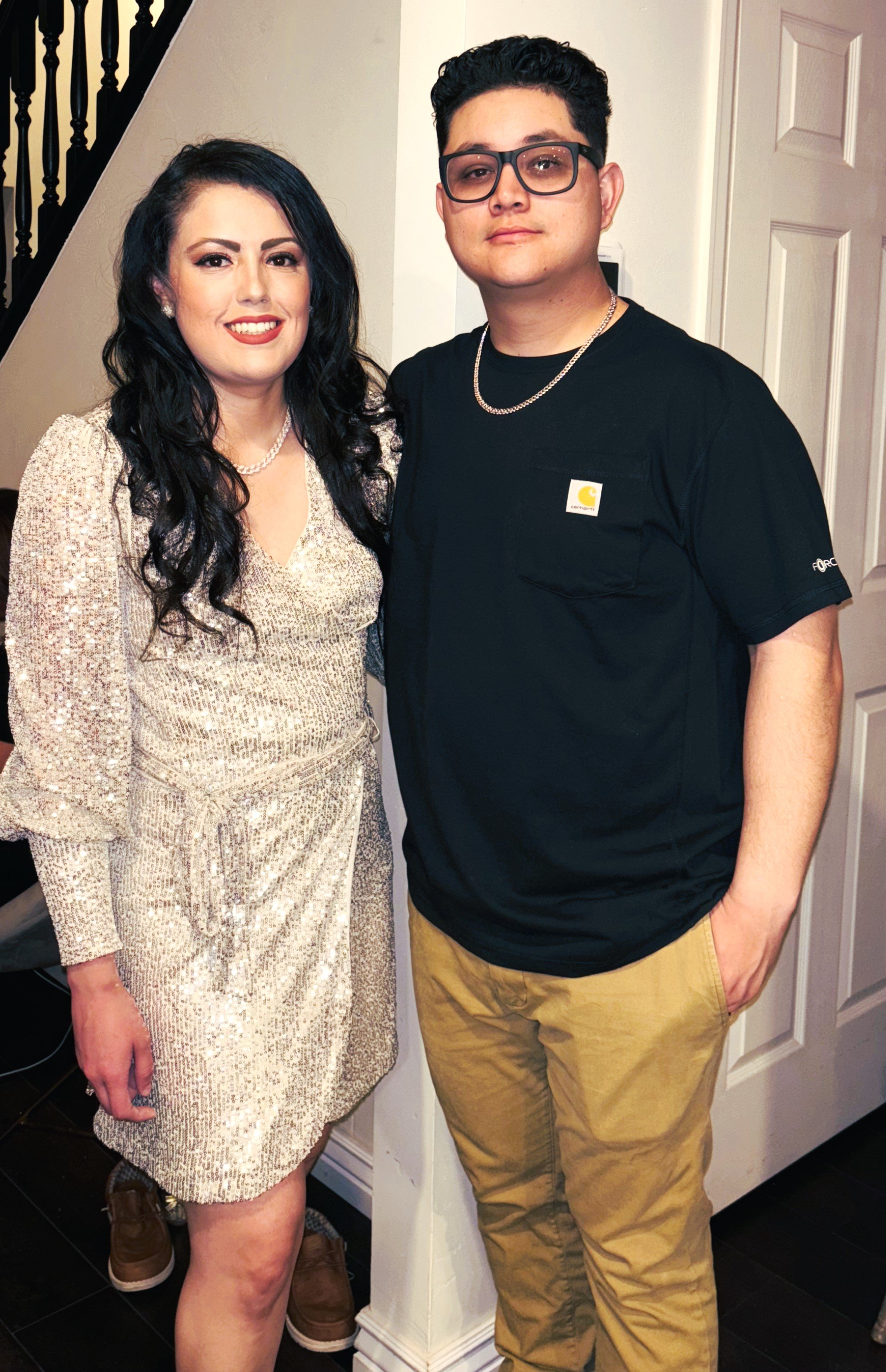 A woman and a young man standing indoors, smiling for the camera. The woman is wearing a shiny, gold-colored dress with a pearl necklace, and has long, dark, wavy hair and makeup. The young man is wearing glasses, a black T-shirt, a gold chain, and beige pants. They are standing next to a white door and a staircase with dark wood steps.